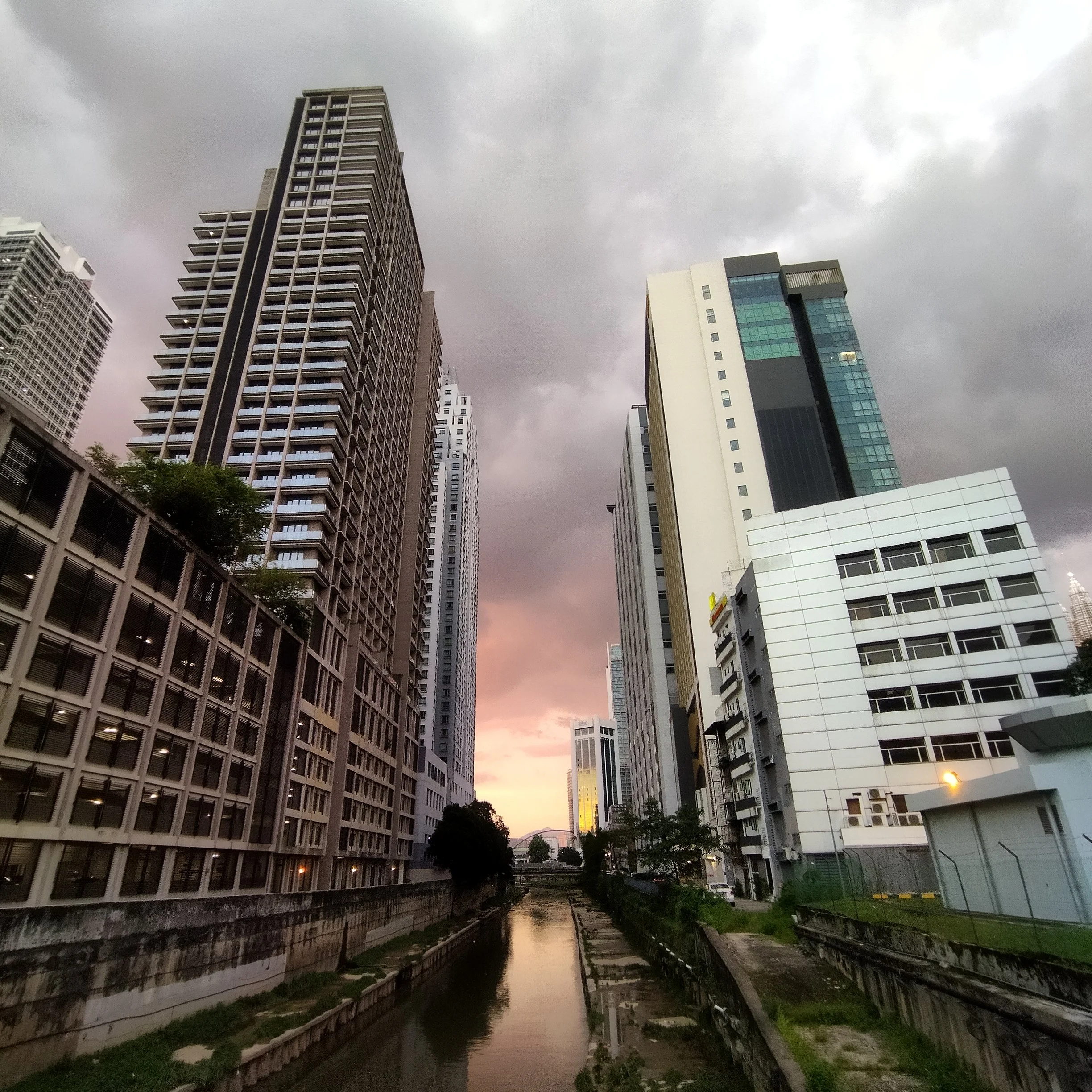 Skyline of tall modern buildings along a small waterway during sunset with dark clouds overhead.