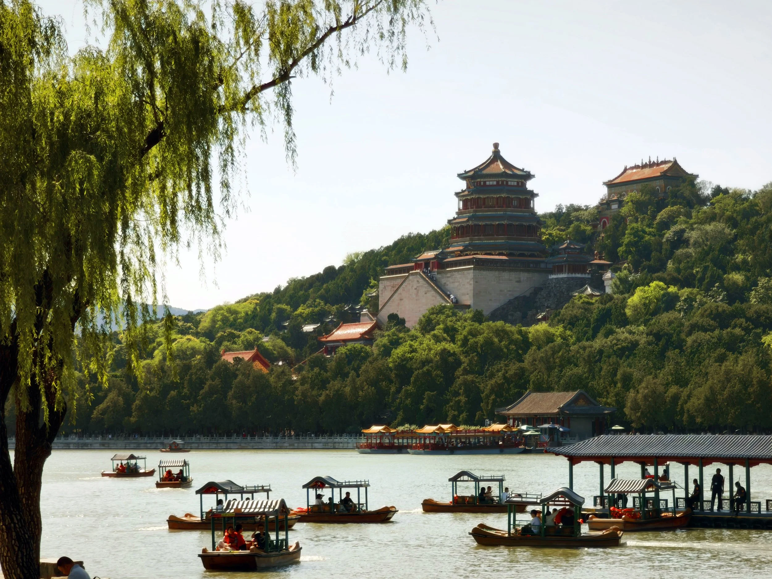 Boats on a lake with a traditional Chinese palace on a hill in the background, lush green trees surrounding the area, and people on the boats