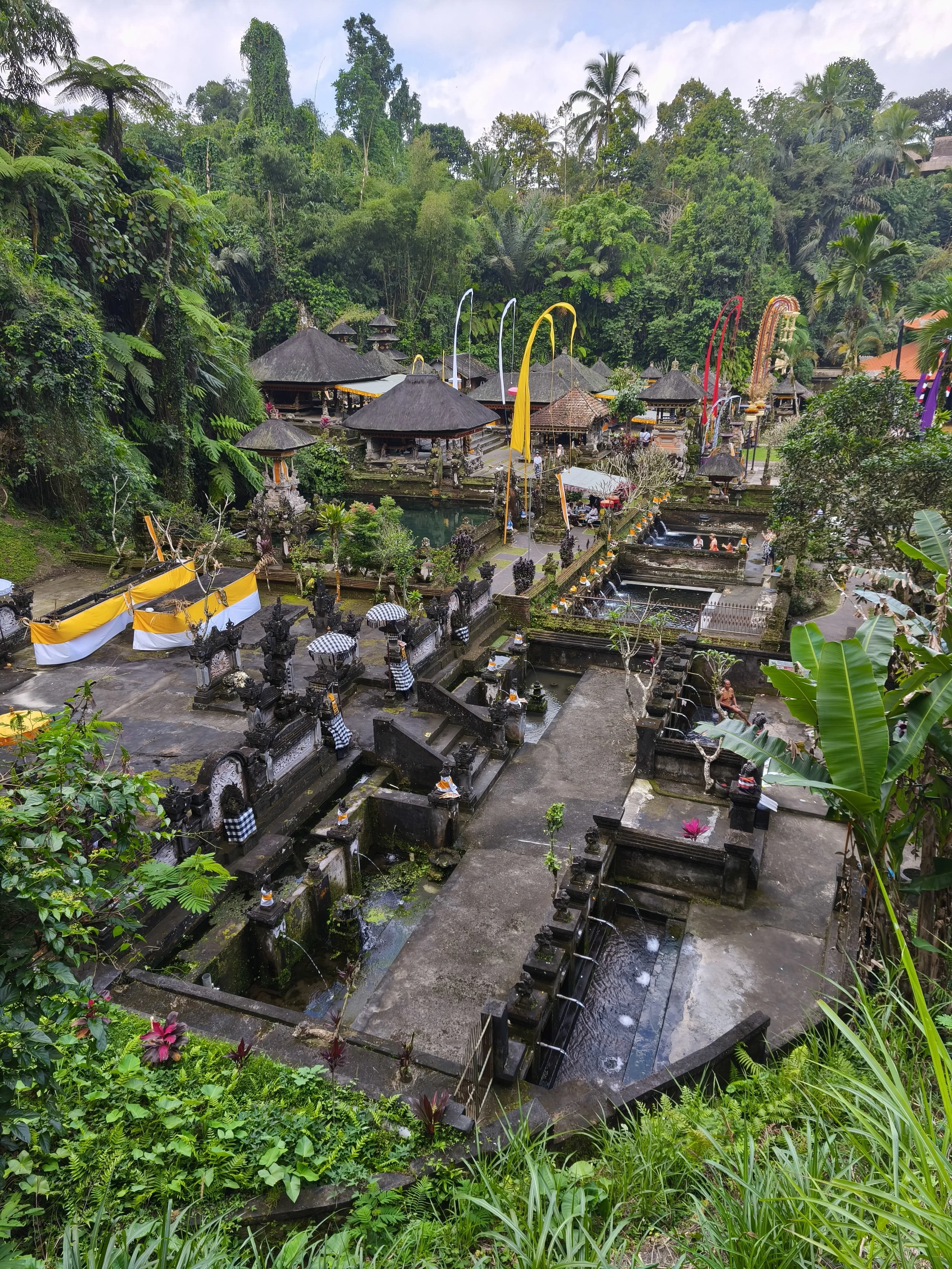 A serene Balinese temple surrounded by lush green tropical jungle, with small pavilions, decorative flags, water features, stone statues, and people visiting.