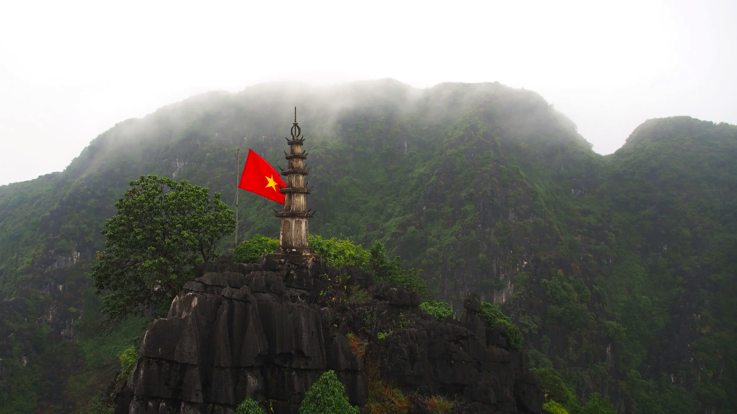 A pagoda-shaped stone monument with a red Vietnam flag featuring a yellow star, situated on a rocky hilltop surrounded by lush greenery and mountains shrouded in mist.