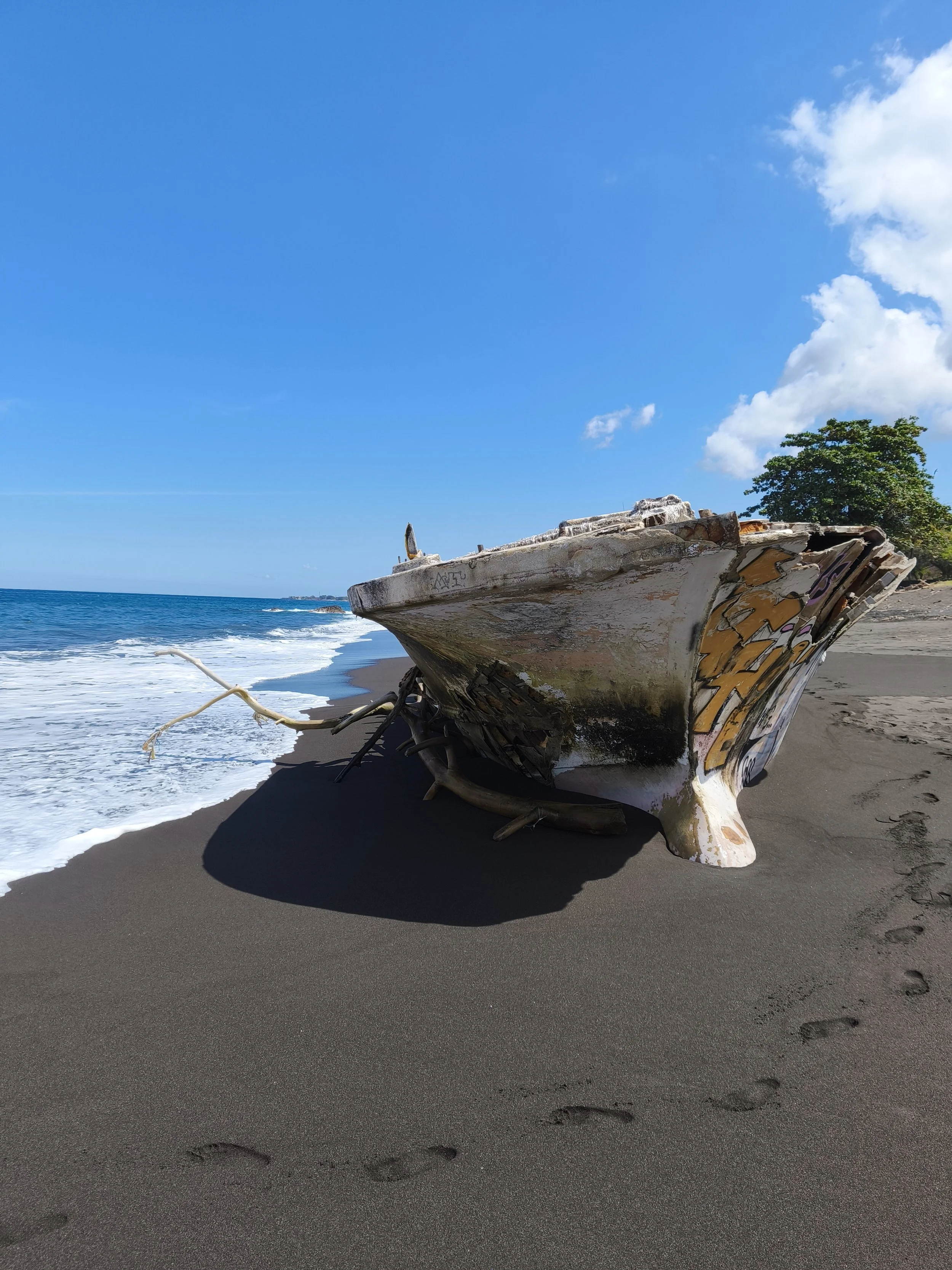 An abandoned, weathered boat on a sandy beach with footprints, under a clear blue sky with some clouds, near the ocean with small waves, and some greenery and trees in the background.