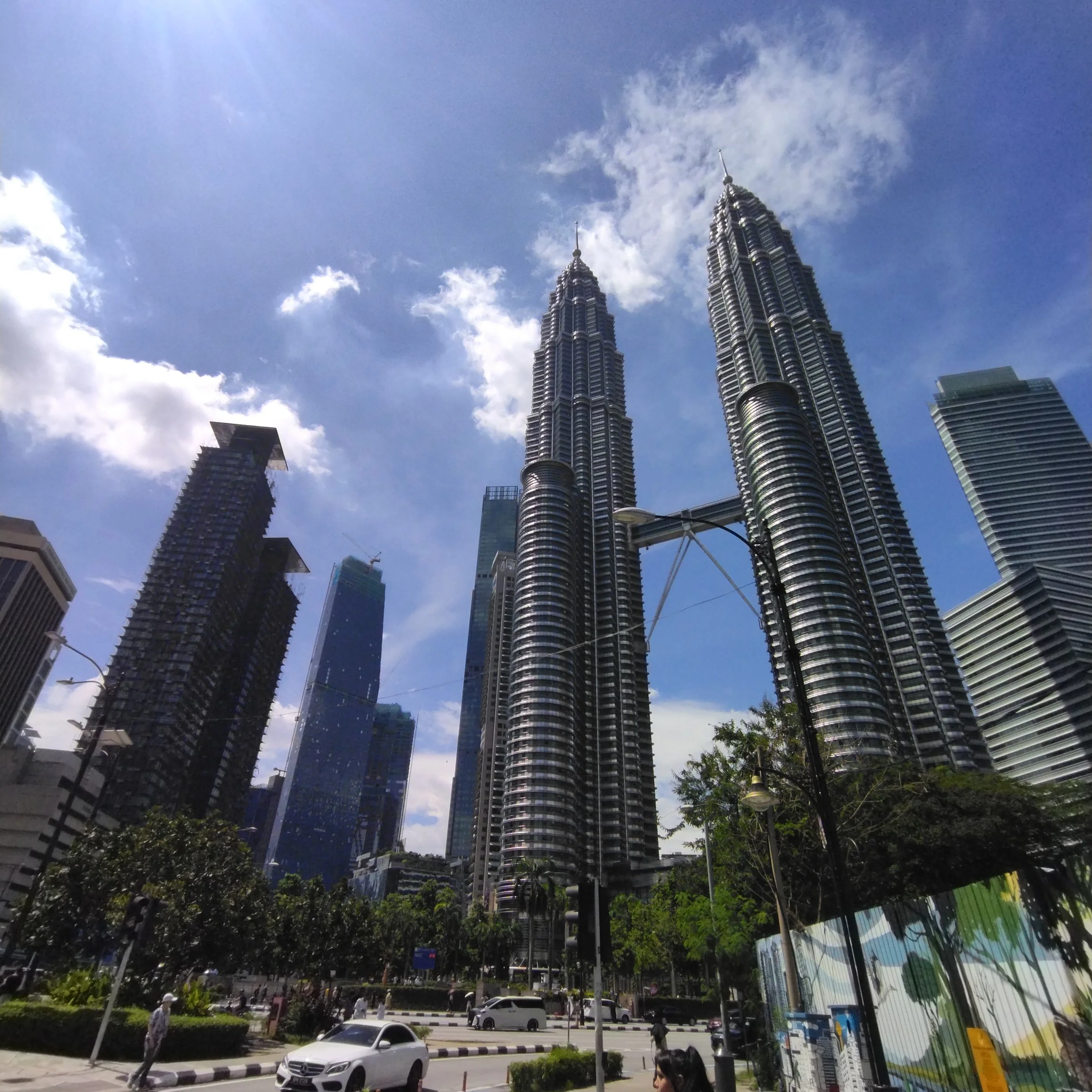 Tall skyscrapers, including the Petronas Towers in Kuala Lumpur, under a partly cloudy sky, with a busy street and trees at the base.