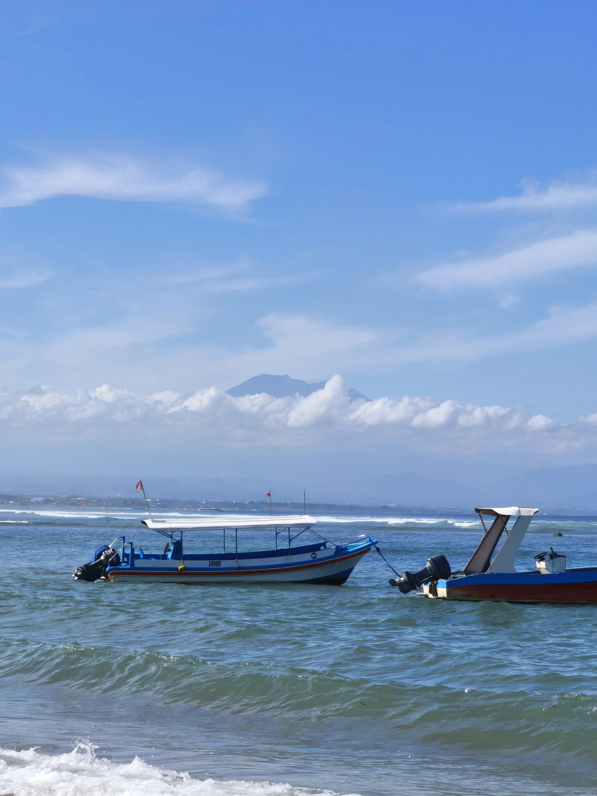 Két kis hajó úszik az óceánon a part közelében, hegyekkel és részben felhős éggel a háttérben.