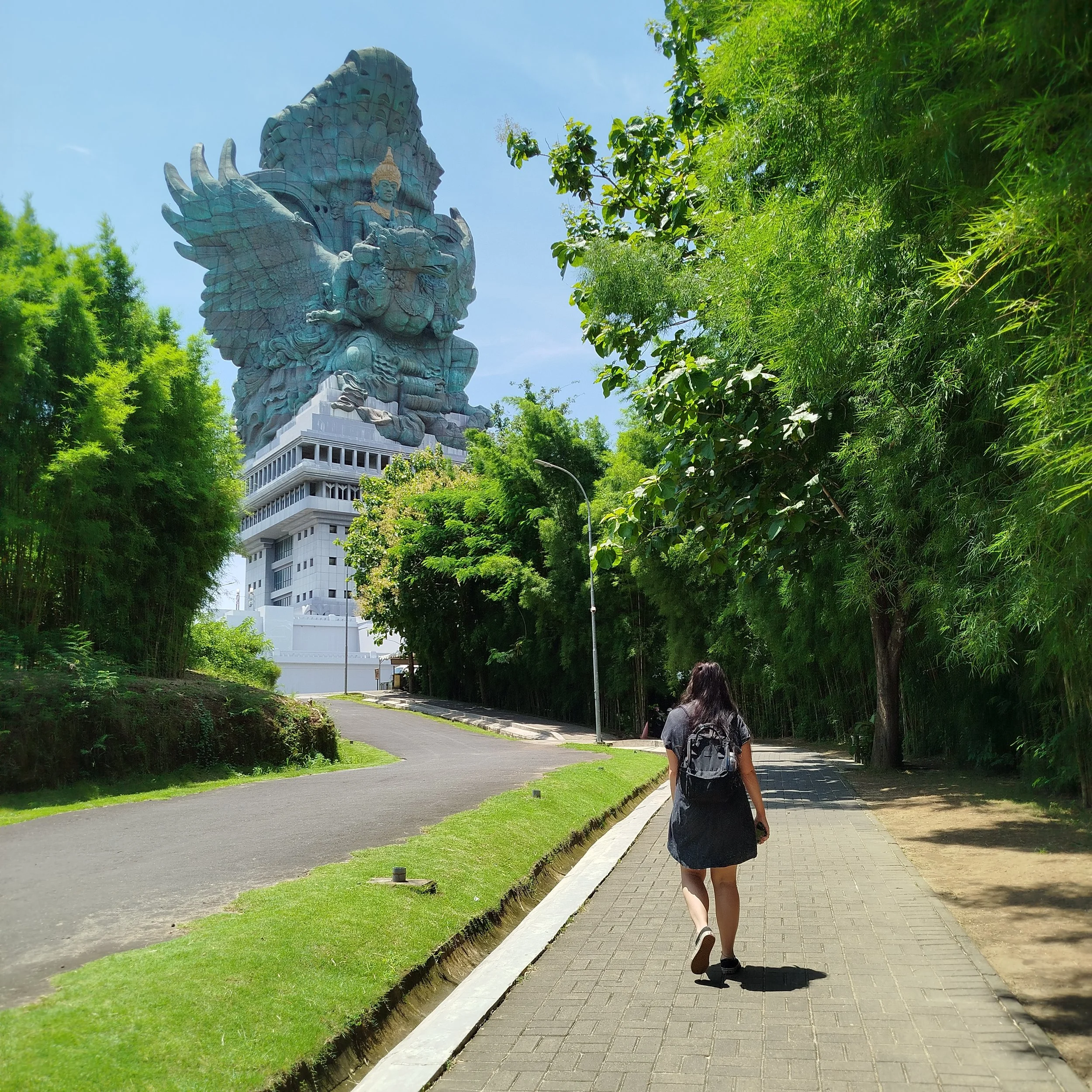 A woman with a backpack walking on a sidewalk surrounded by trees, with a large green sculpture of a mythological creature with wings and a human figure on top in the background.