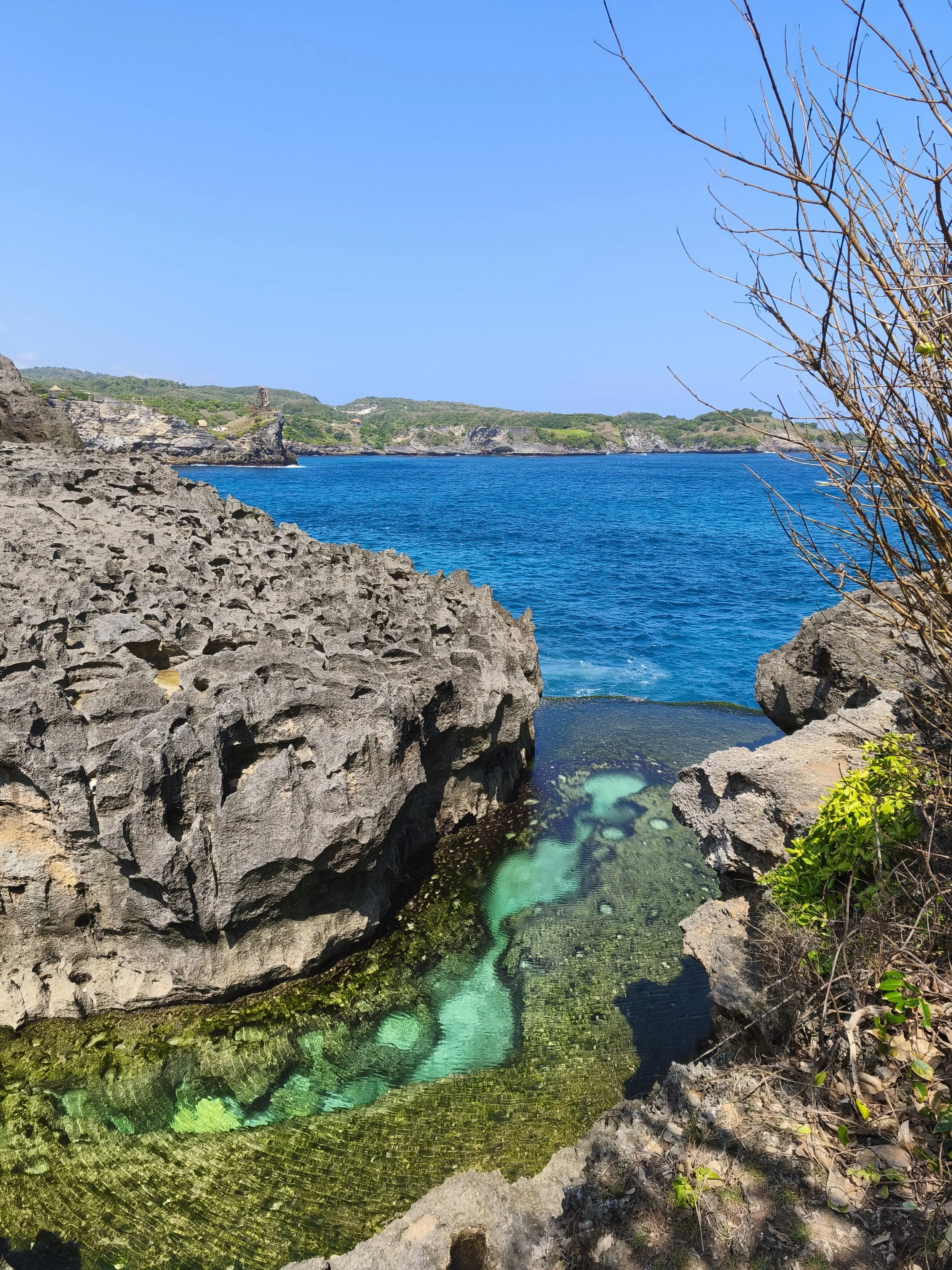 Rocky coastline with a small clear pool of water in the foreground, blue ocean and green cliffs in the background, and a bright blue sky.