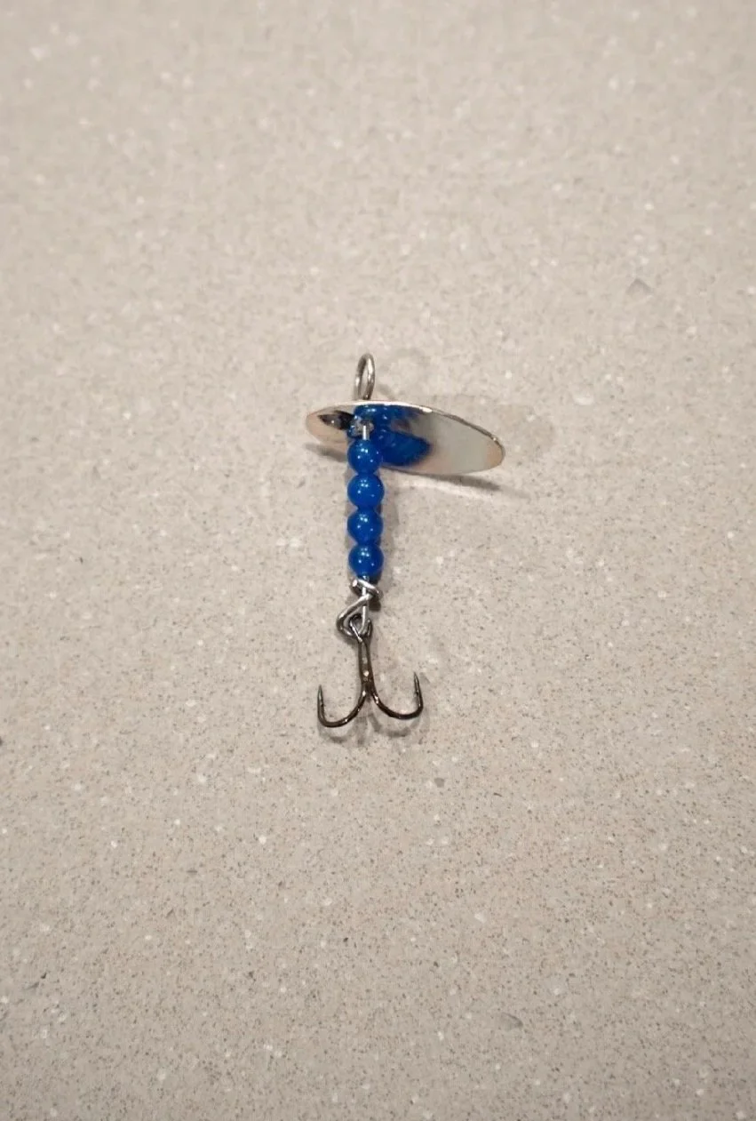 A fishing hook pendant with blue beads and a small fishhook charm, attached to a silver ring on a beige surface.