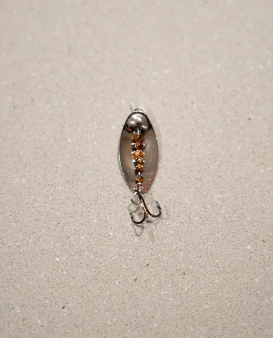A fishing lure with a small metal ball, a row of small beads, and two hooks, placed on a beige surface.