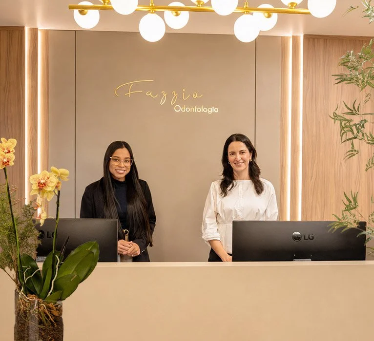 Dra. Silvia e recepcionistas sorridentes na recepção da Clínica Fazzio, em ambiente com decoração moderna e elementos naturais, refletindo o cuidado com o bem-estar desde a chegada dos pacientes em Alphaville.