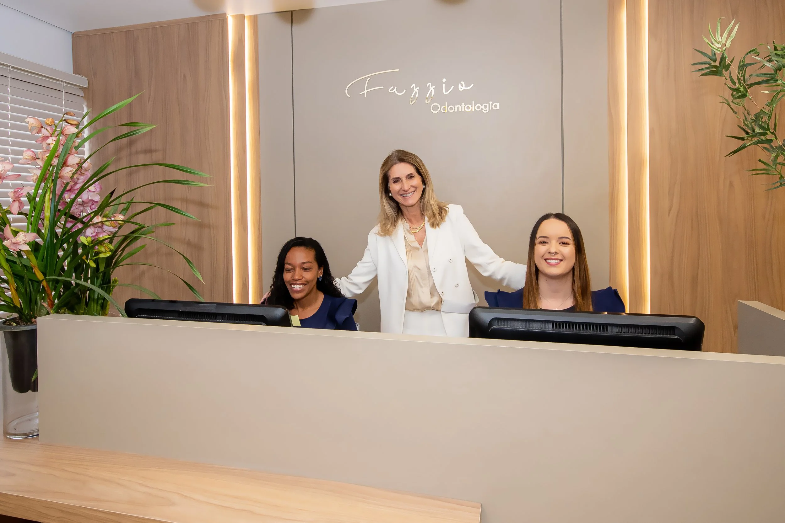 Dra. Silvia e recepcionistas sorridentes na recepção da Clínica Fazzio, em ambiente com decoração moderna e elementos naturais, refletindo o cuidado com o bem-estar desde a chegada dos pacientes em Alphaville.