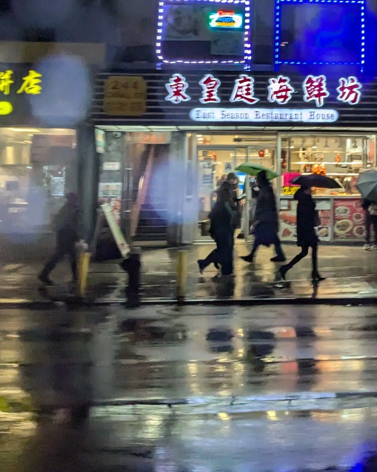 Nighttime street scene with people walking under umbrellas, reflecting wet pavement, in front of a restaurant with illuminated sign and Asian characters.