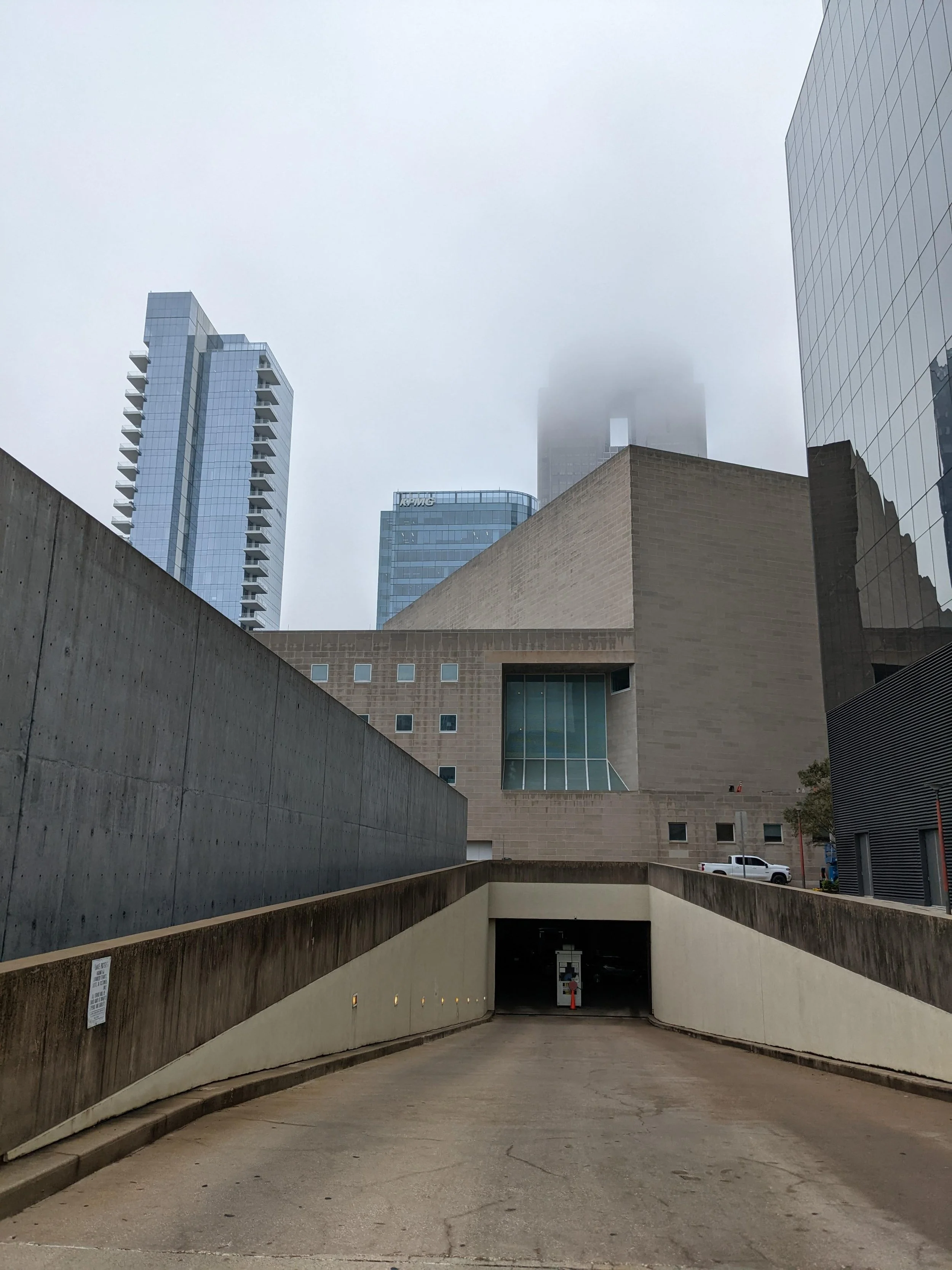 The image depicts a modern urban scene with tall buildings, some partially obscured by fog, and a parking garage entrance in the foreground.