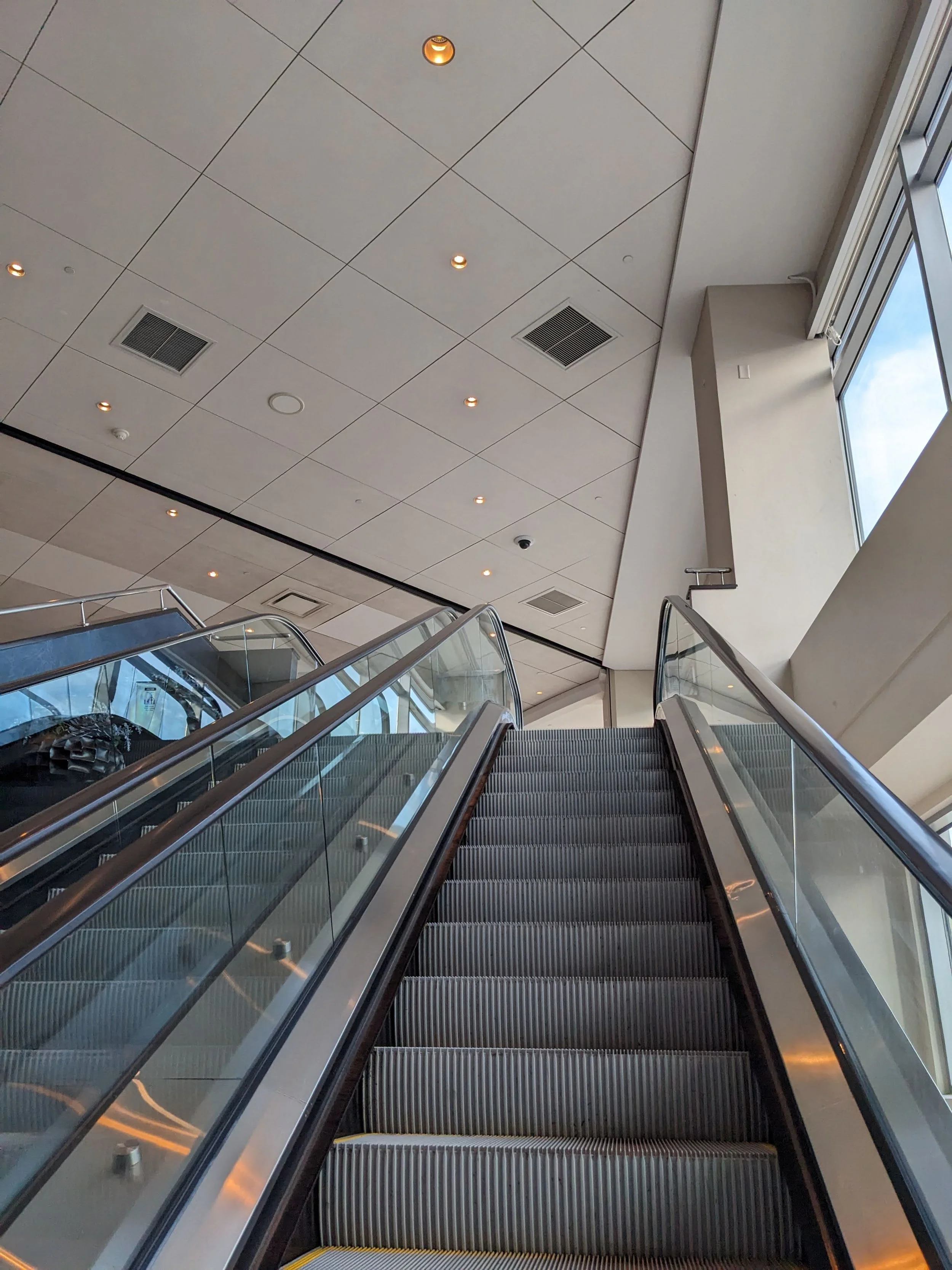 Escalator in a modern building with a white ceiling, small round recessed lights, and air vents, leading upward near a large window showing sky and clouds.