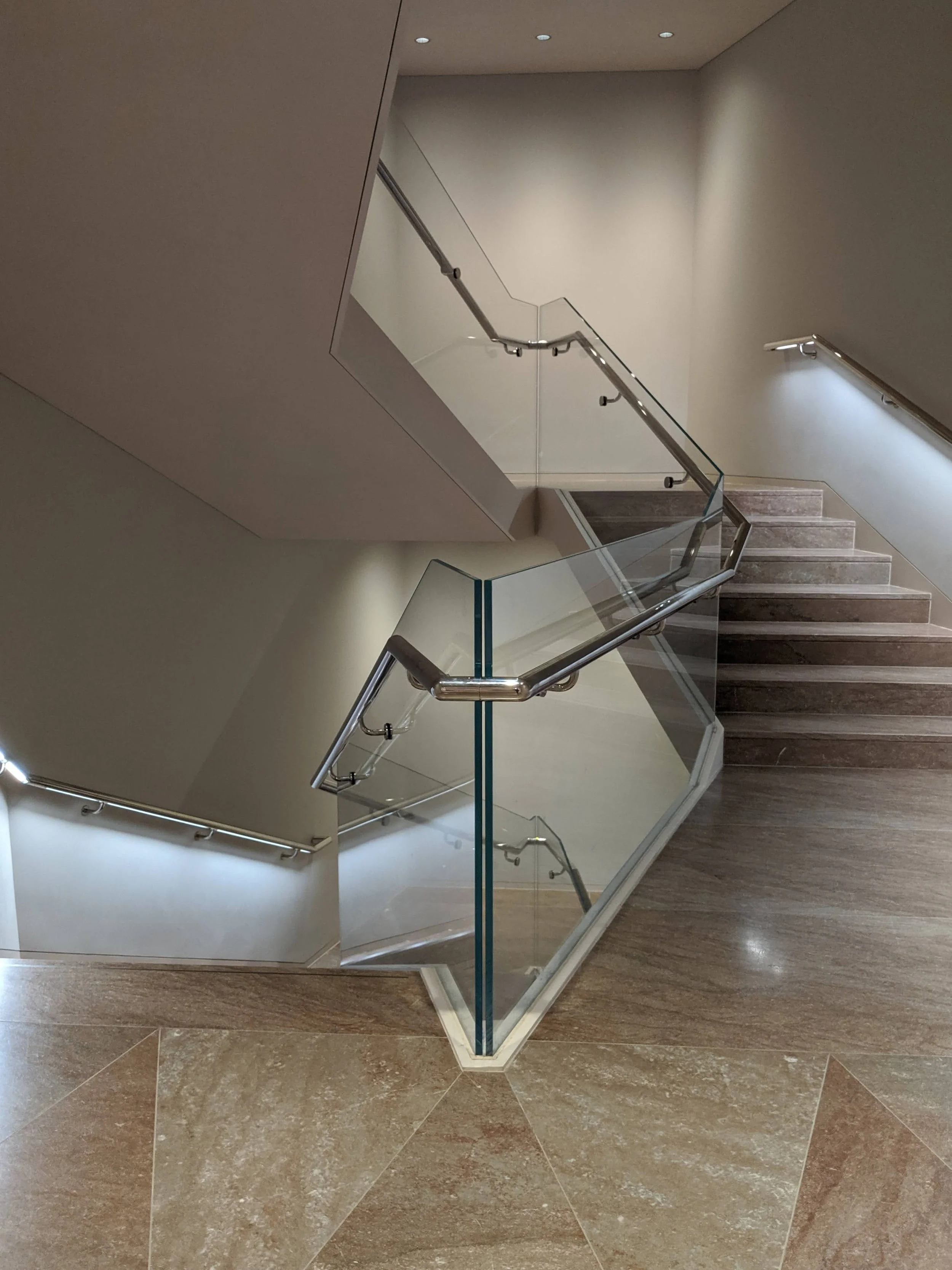 Interior shot of a modern staircase with glass and metal handrails, beige tiled steps, and a wall with small ceiling lights above.