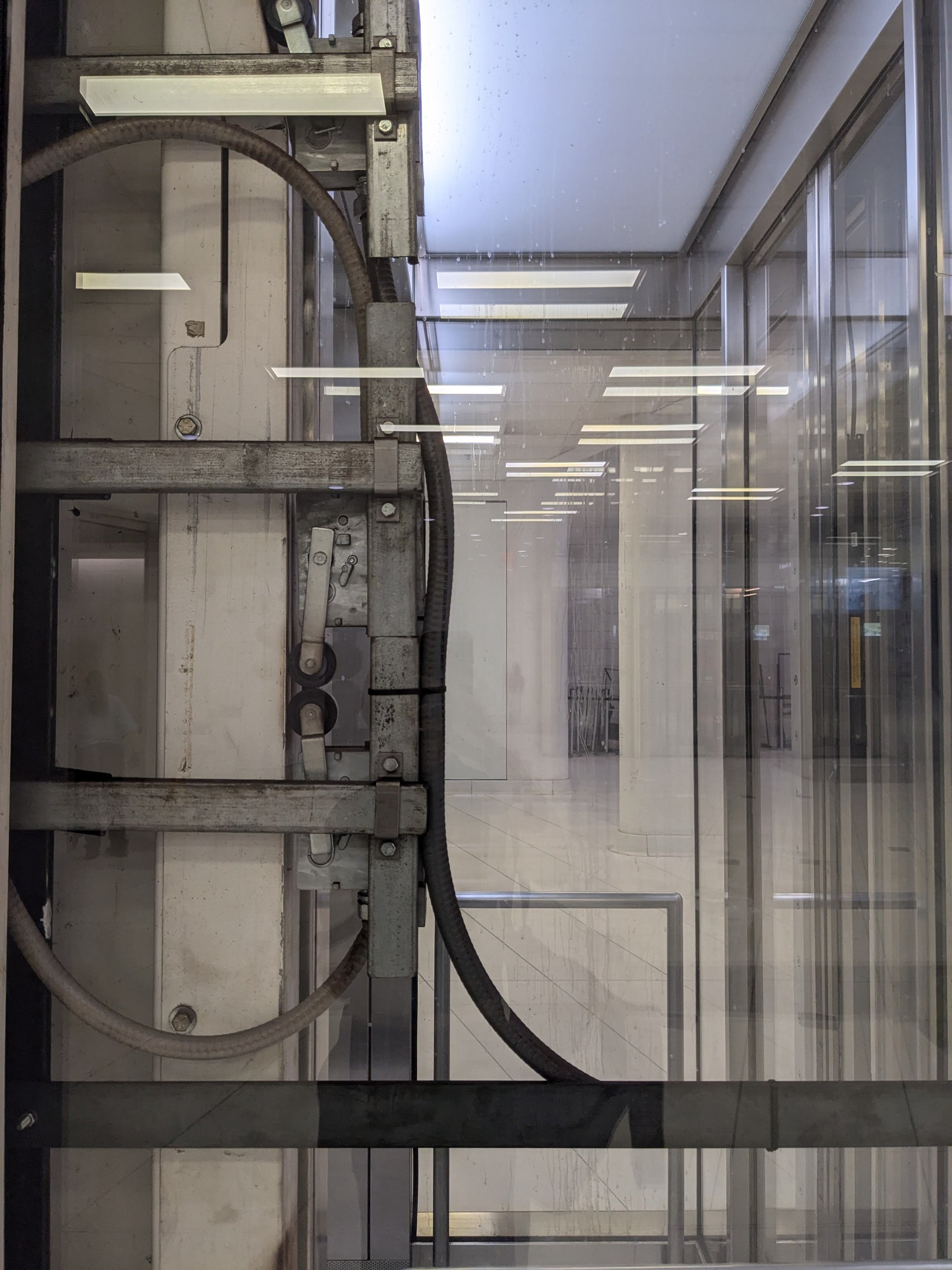 Close-up of industrial mechanical parts and cables inside a glass-enclosed area with reflections of fluorescent lights.