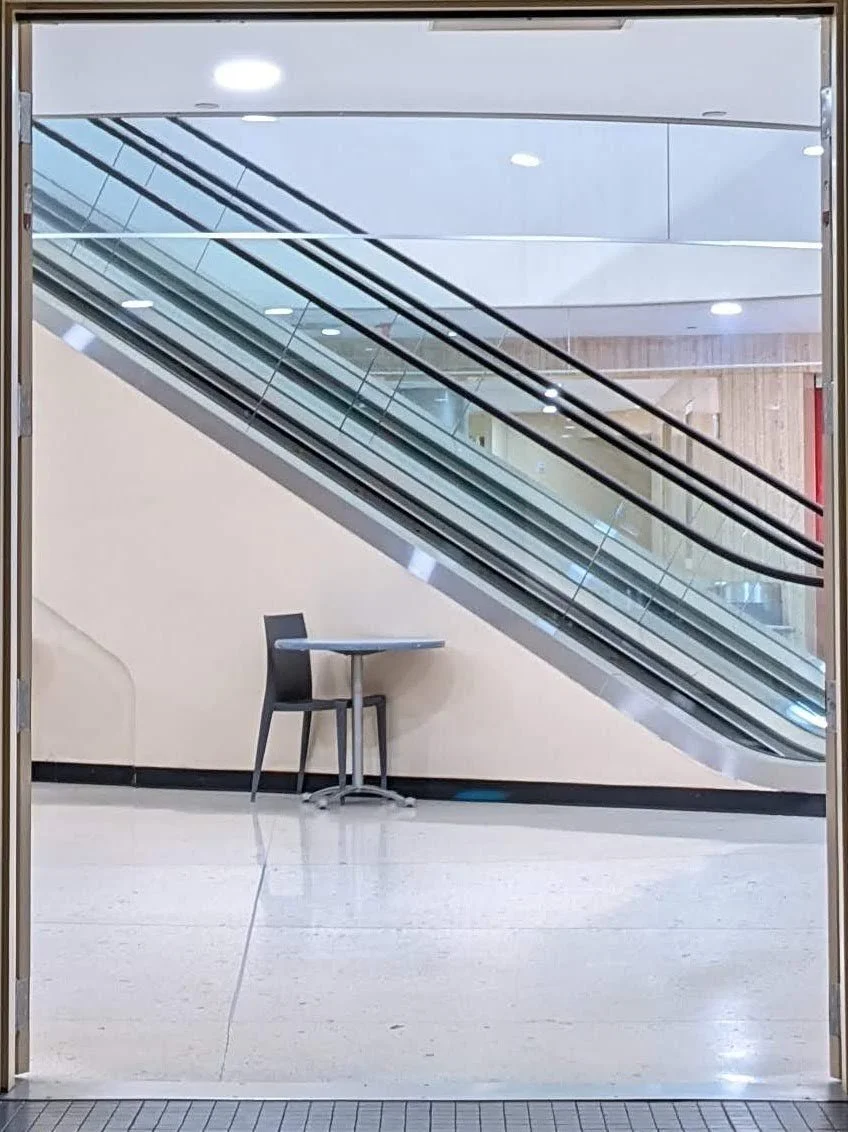A glass door reflects an enclosed area in a mall with an escalator and a small table with a chair underneath it.