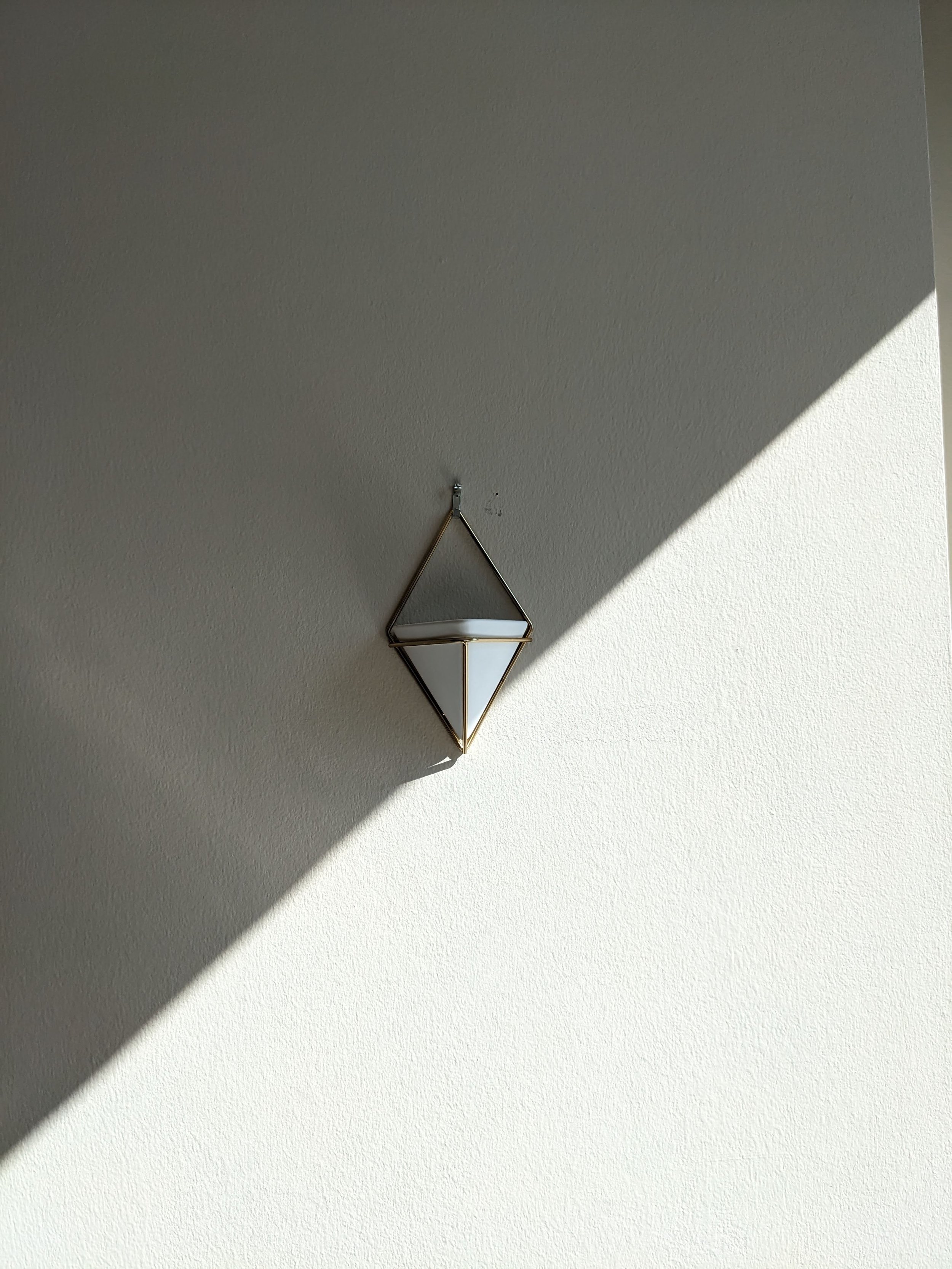 A wall-mounted geometric mirror with a triangular frame, hanging on a textured wall with sunlight casting a shadow.