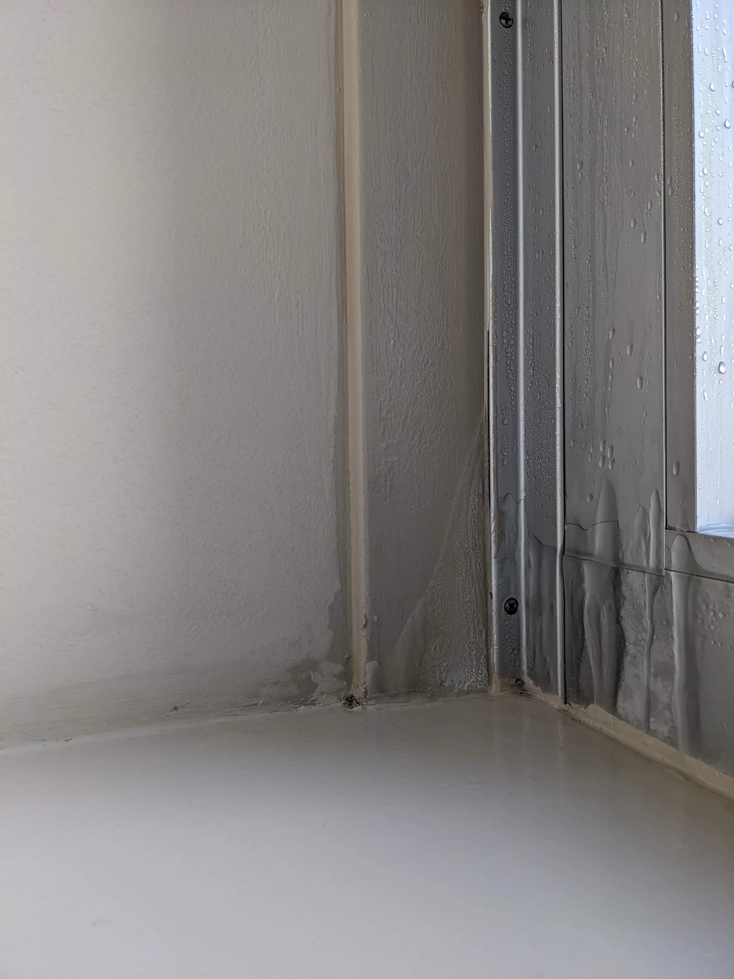 Close-up image of a section of a wall with a window frame, showing exposed drywall and drywall tape, with some water droplets on the window surface.