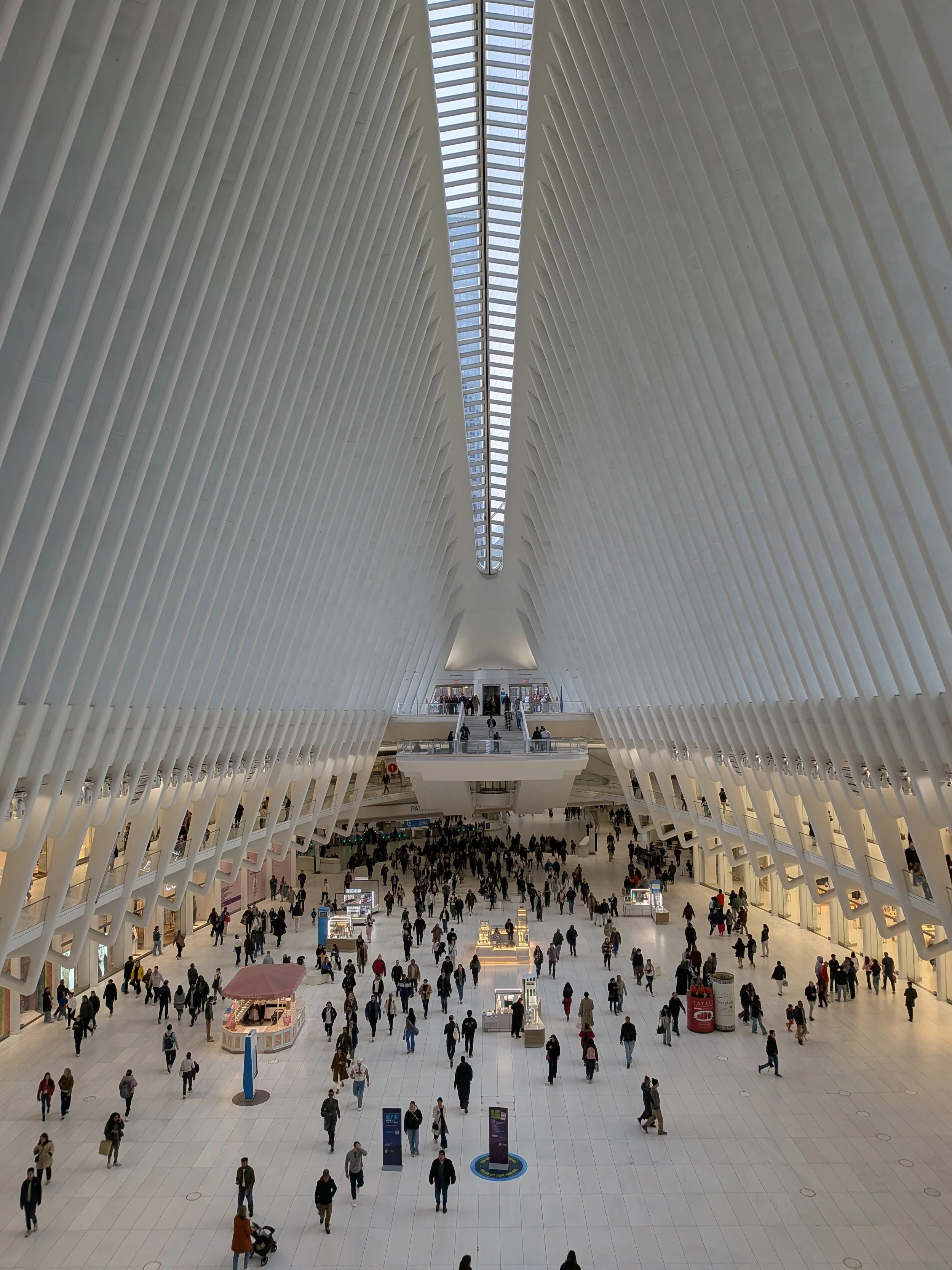 A busy airport terminal with high, ribbed white architectural walls, a skylight ceiling, and crowds of people walking around the open space.