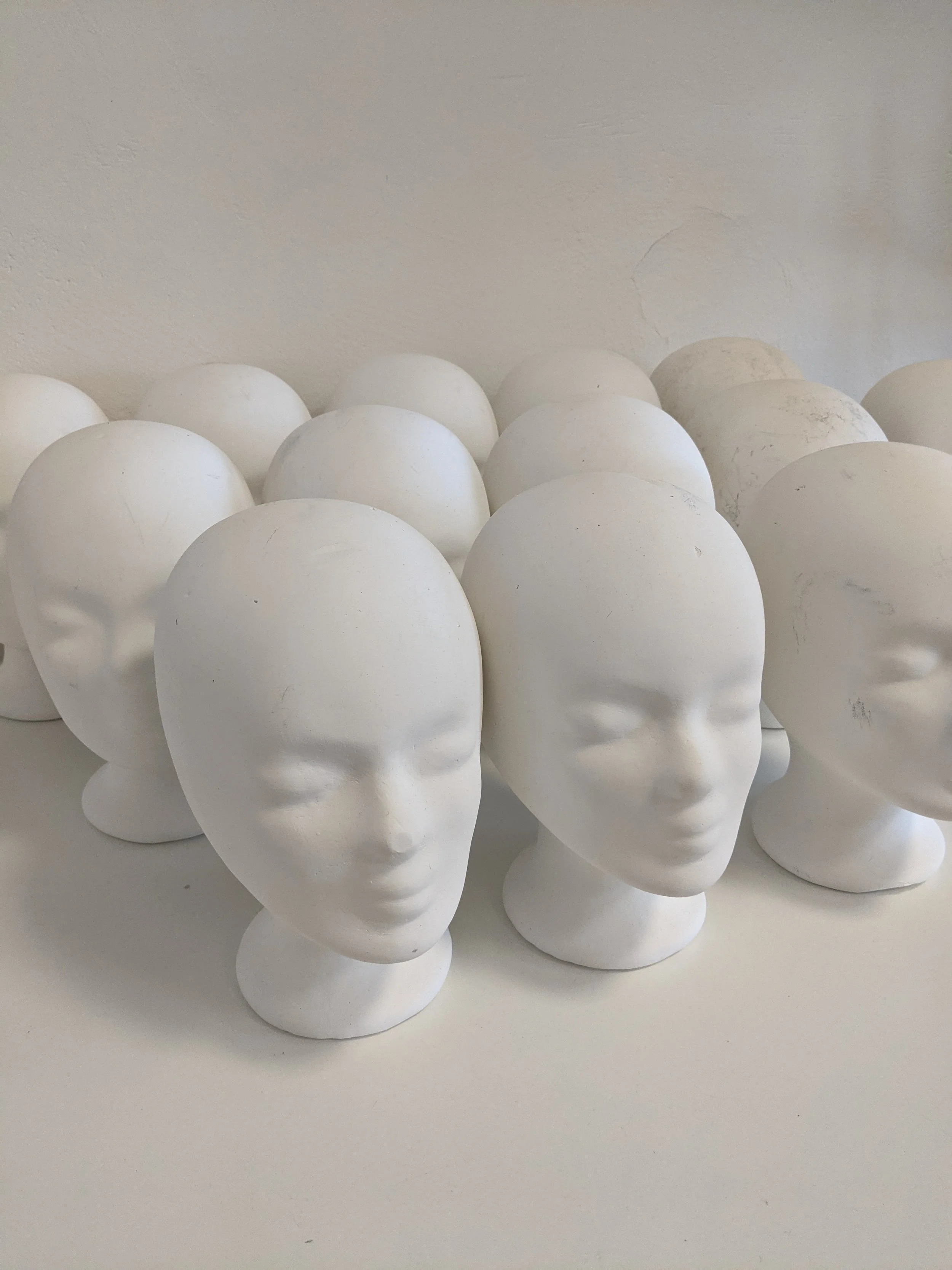 Multiple white ceramic or plaster head sculptures arranged on a plain surface against a plain wall.