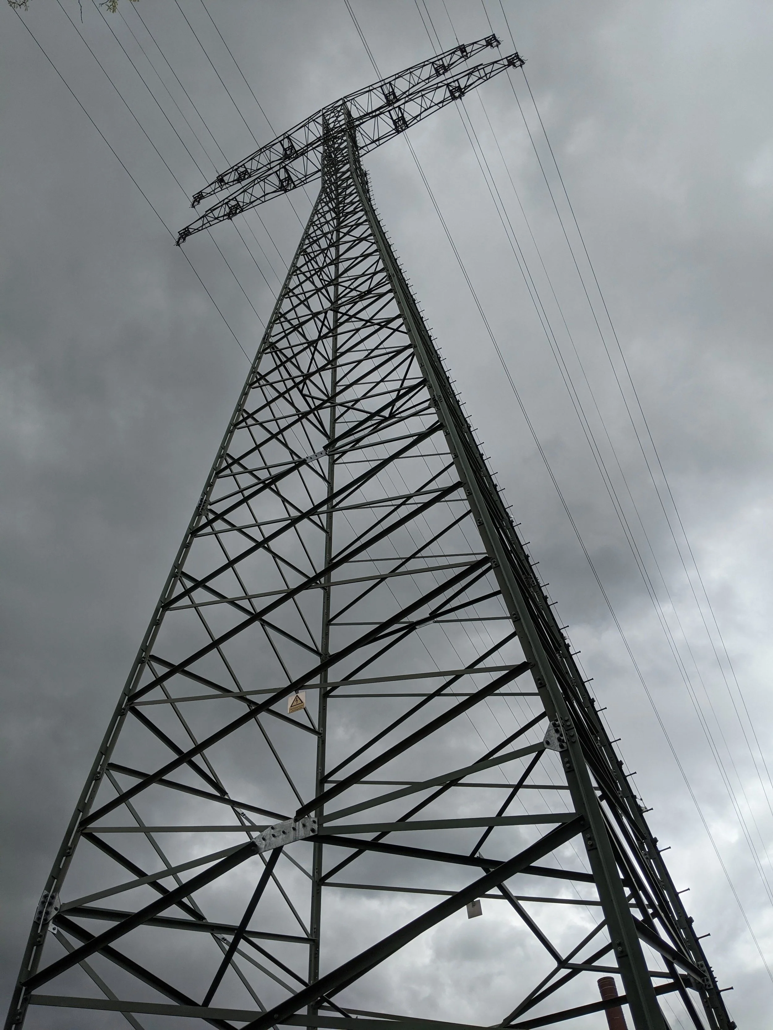 A tall steel power line transmission tower seen from below against a cloudy, gray sky.