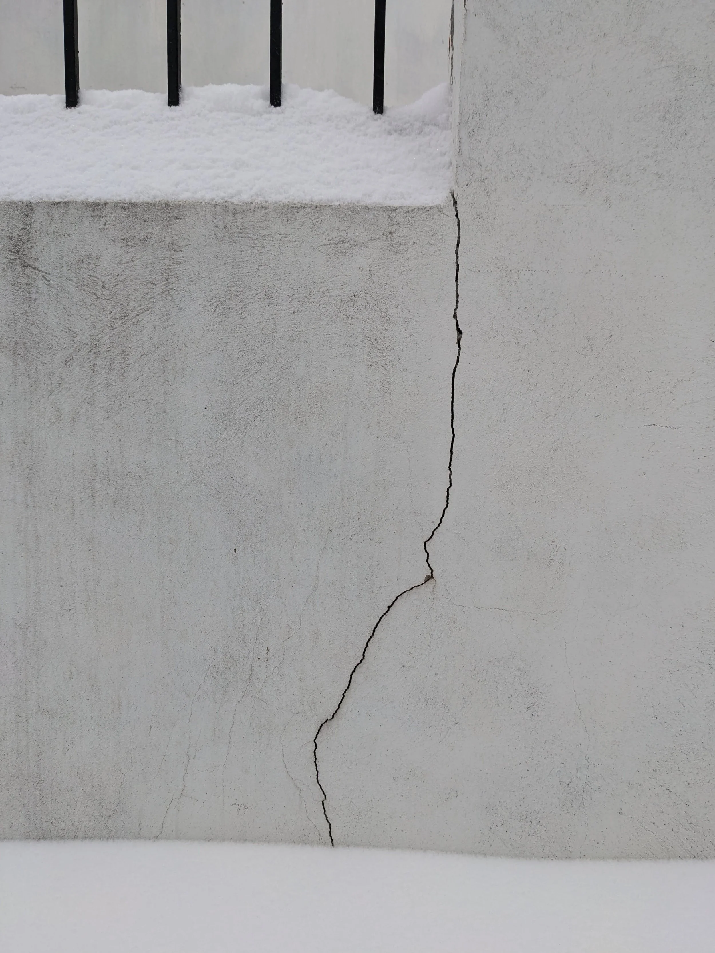 Close-up of a crack in a white wall or concrete surface, with the crack starting at the bottom and extending upward, revealing some snow or ice on an elevated surface above.