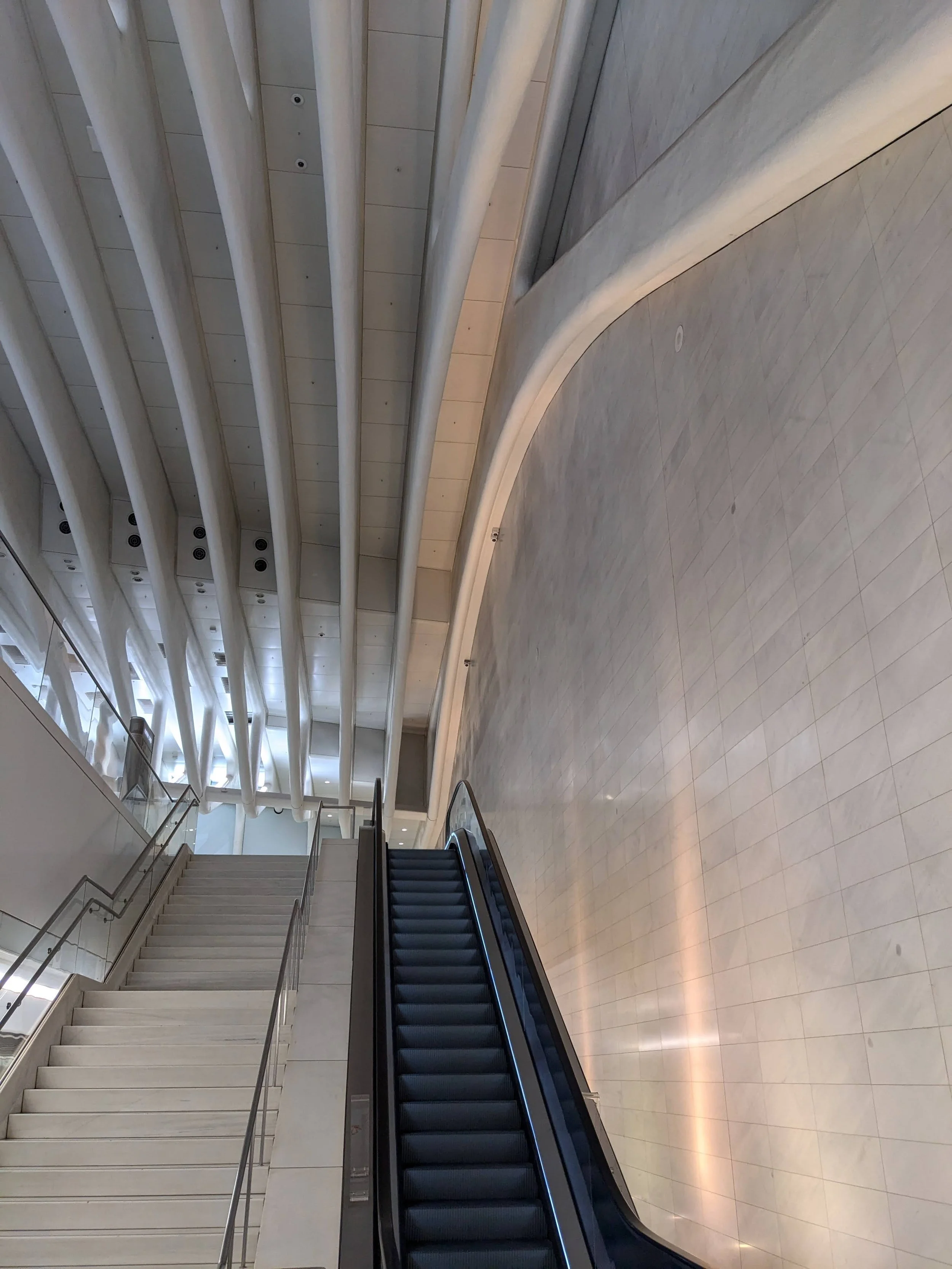 Inside view of a modern shopping mall or airport with a wide open space, marble flooring, curved wall, and an escalator leading upstairs.