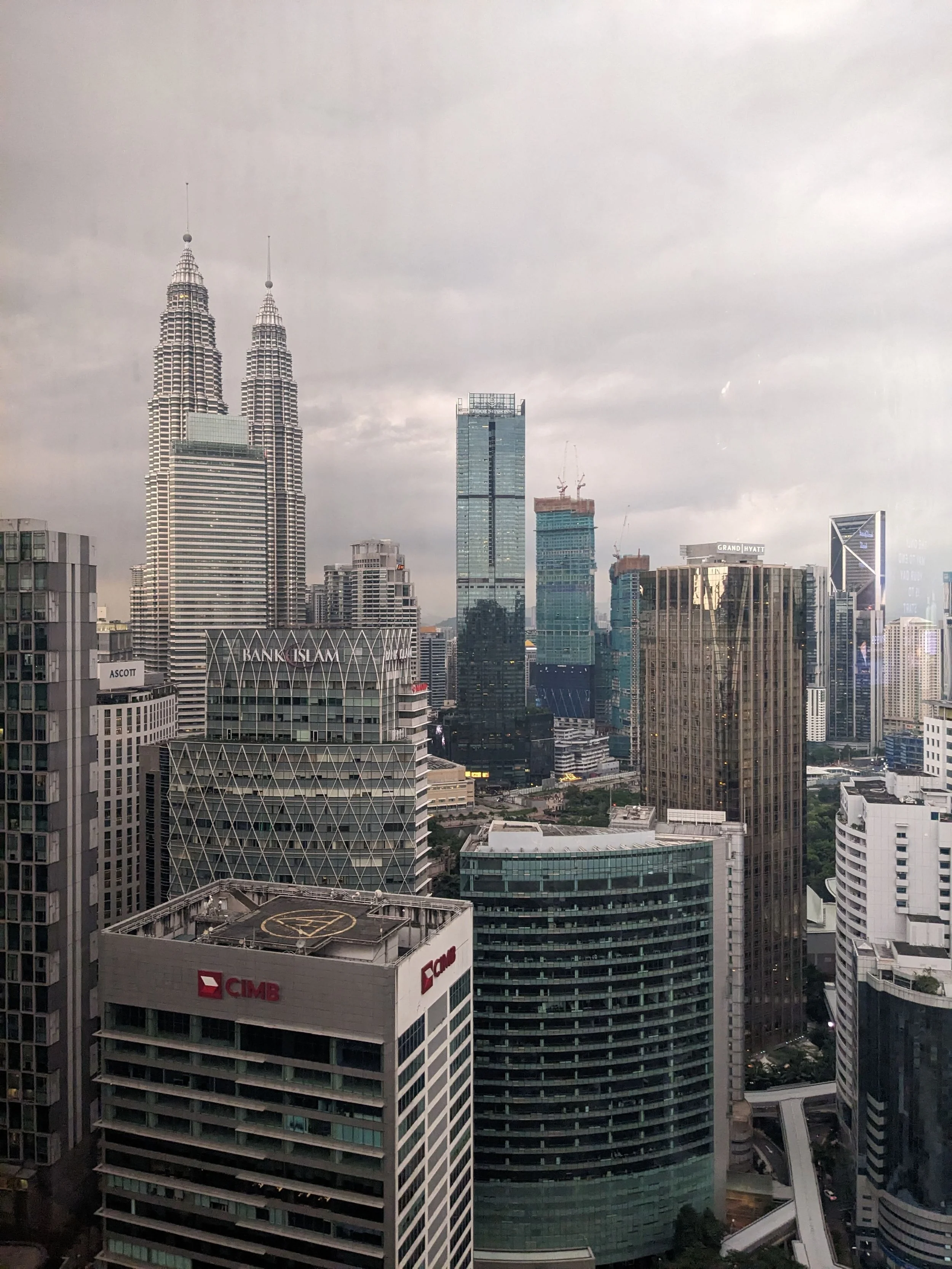 A cityscape featuring skyscrapers including the Petronas Towers, a tall glass building, and other high-rise buildings with a cloudy sky overhead.