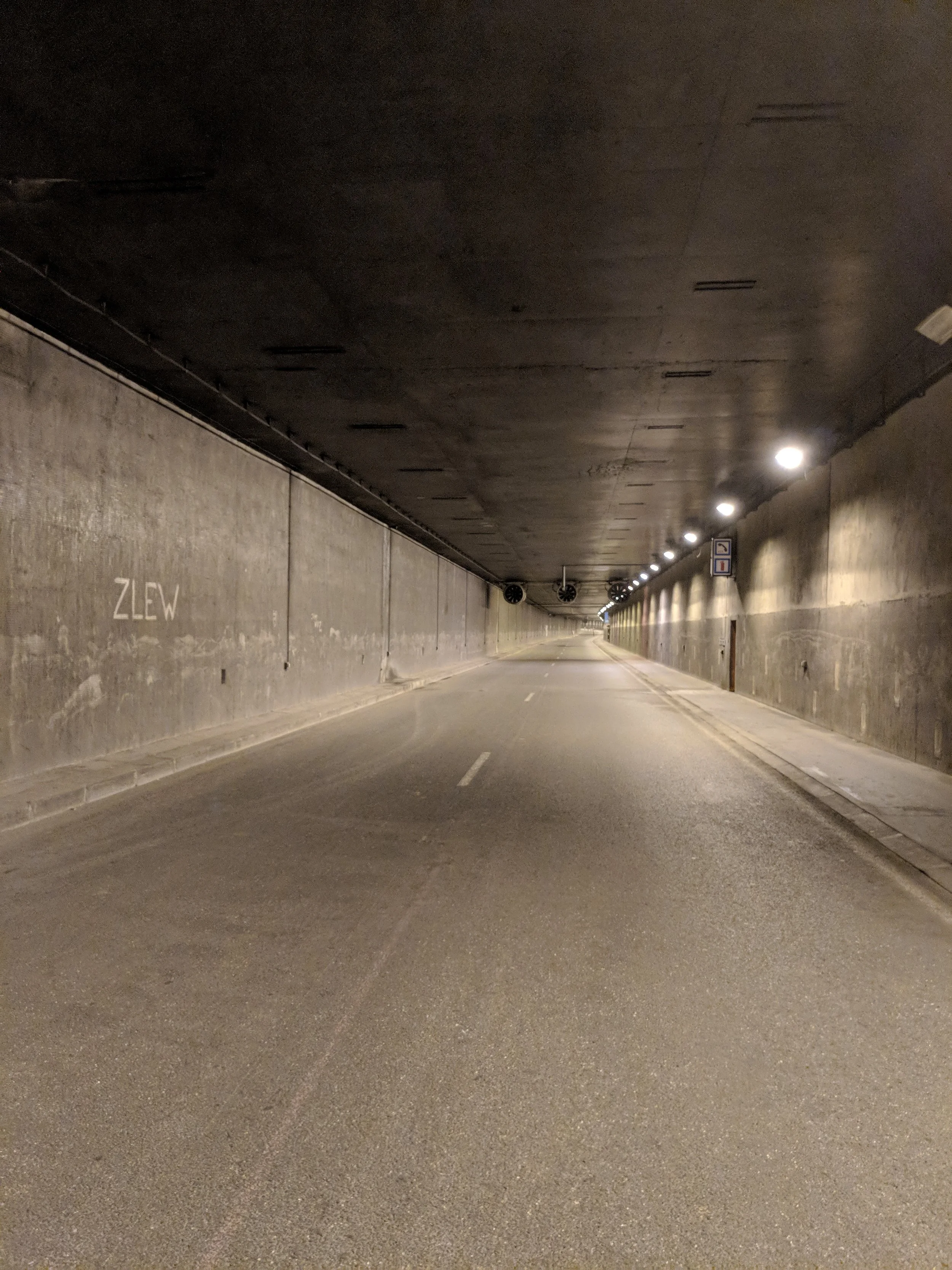 Empty underground tunnel with concrete walls and ceiling. The tunnel has a row of bright ceiling lights on the right side. The road inside the tunnel is marked with white dashed lines and appears clean and well-maintained.