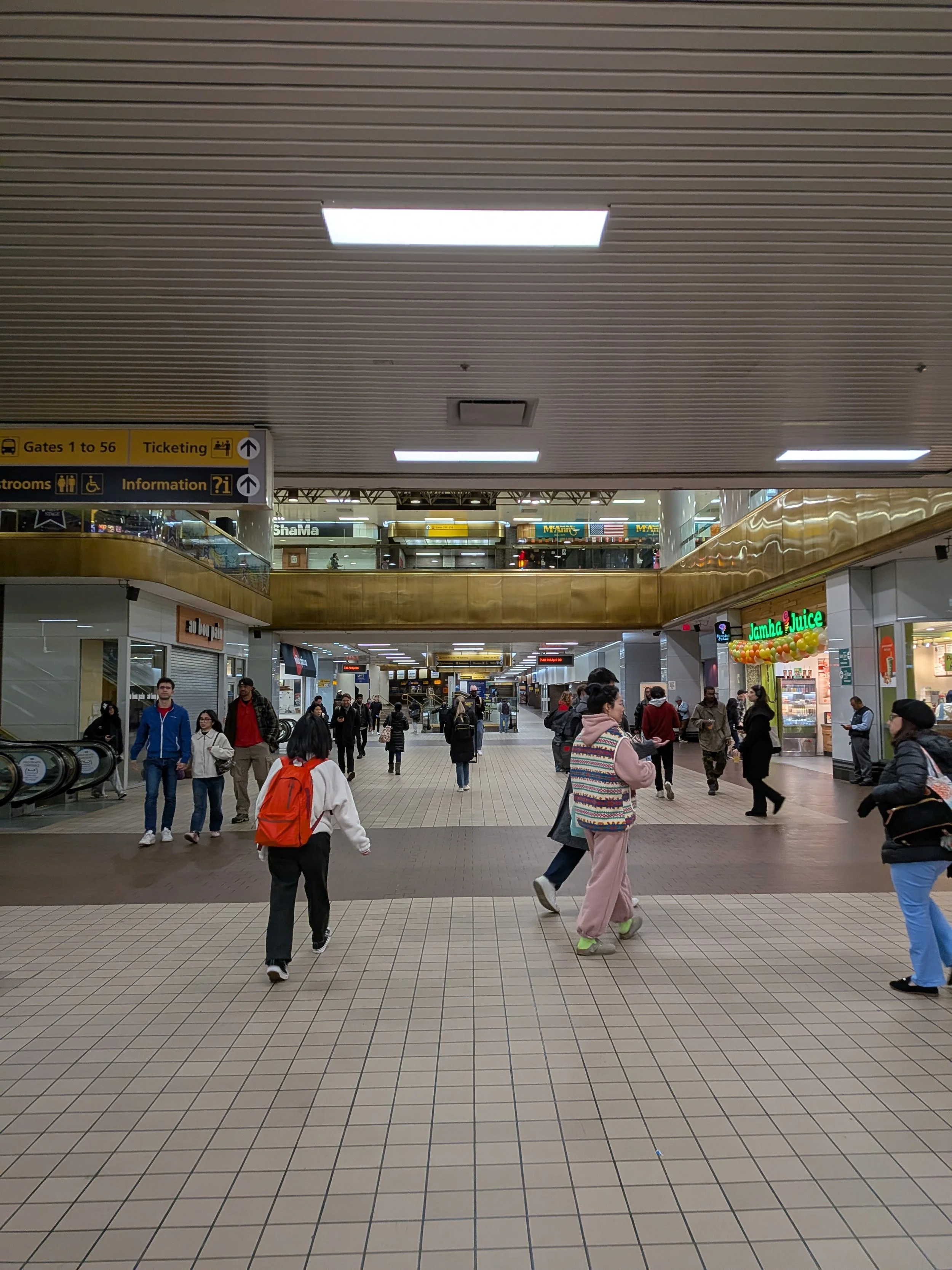People walking in a busy airport terminal with shops and signs overhead including directions to gates, ticketing, restrooms, and information.