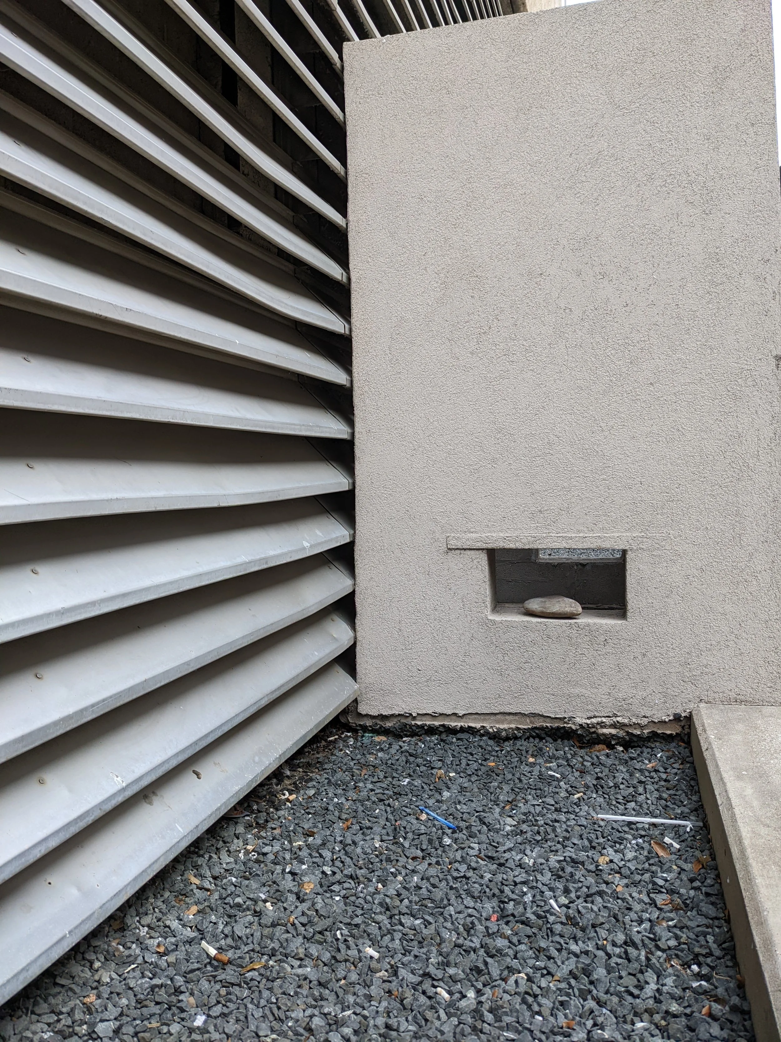 A building exterior with a metal louvered vent on the left, a beige stucco wall on the right, and gravel ground with some trash and debris.