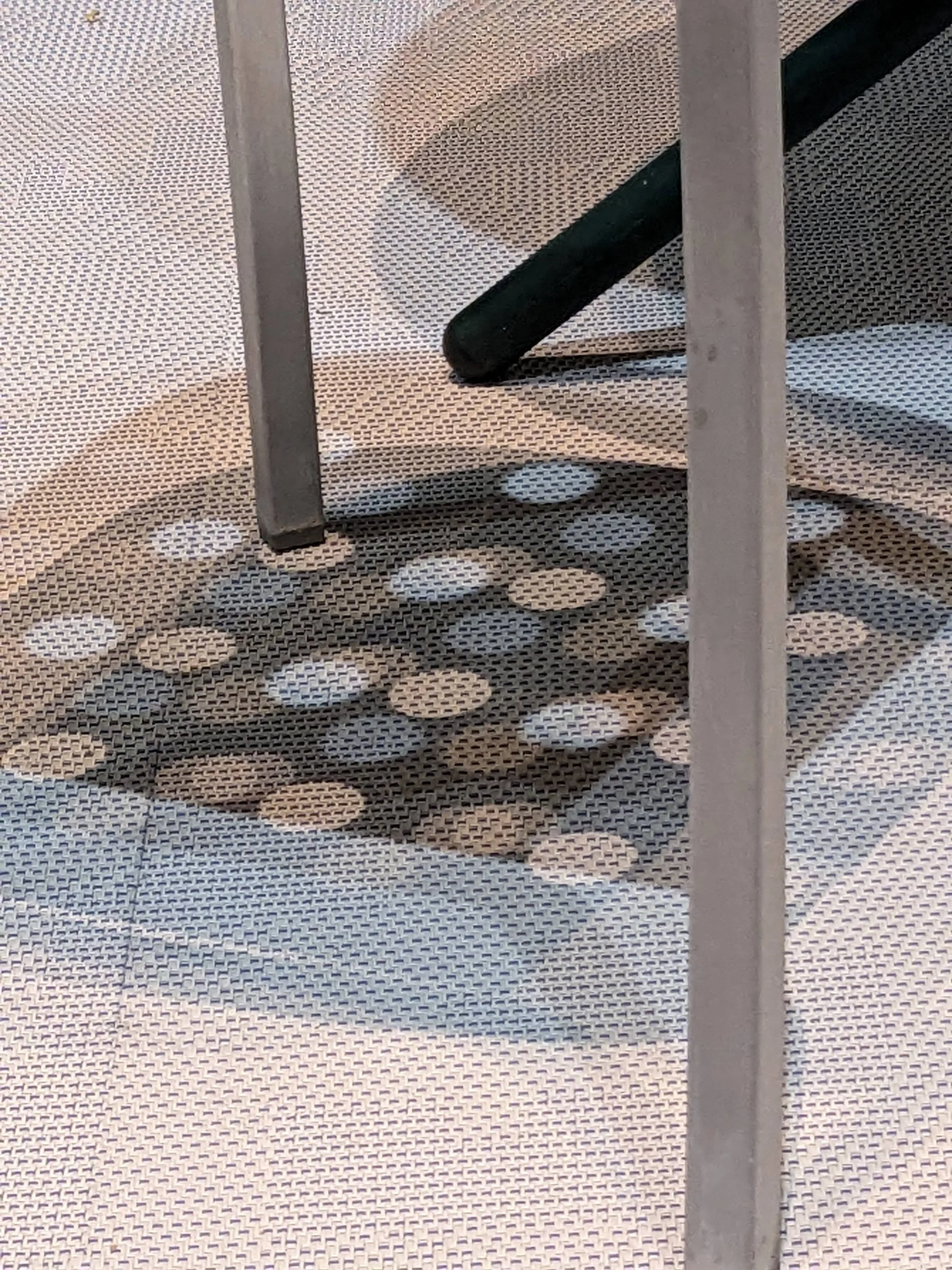 Shadow of a circular metal chair base with round holes projected on a textured light surface, framed by two chair legs.