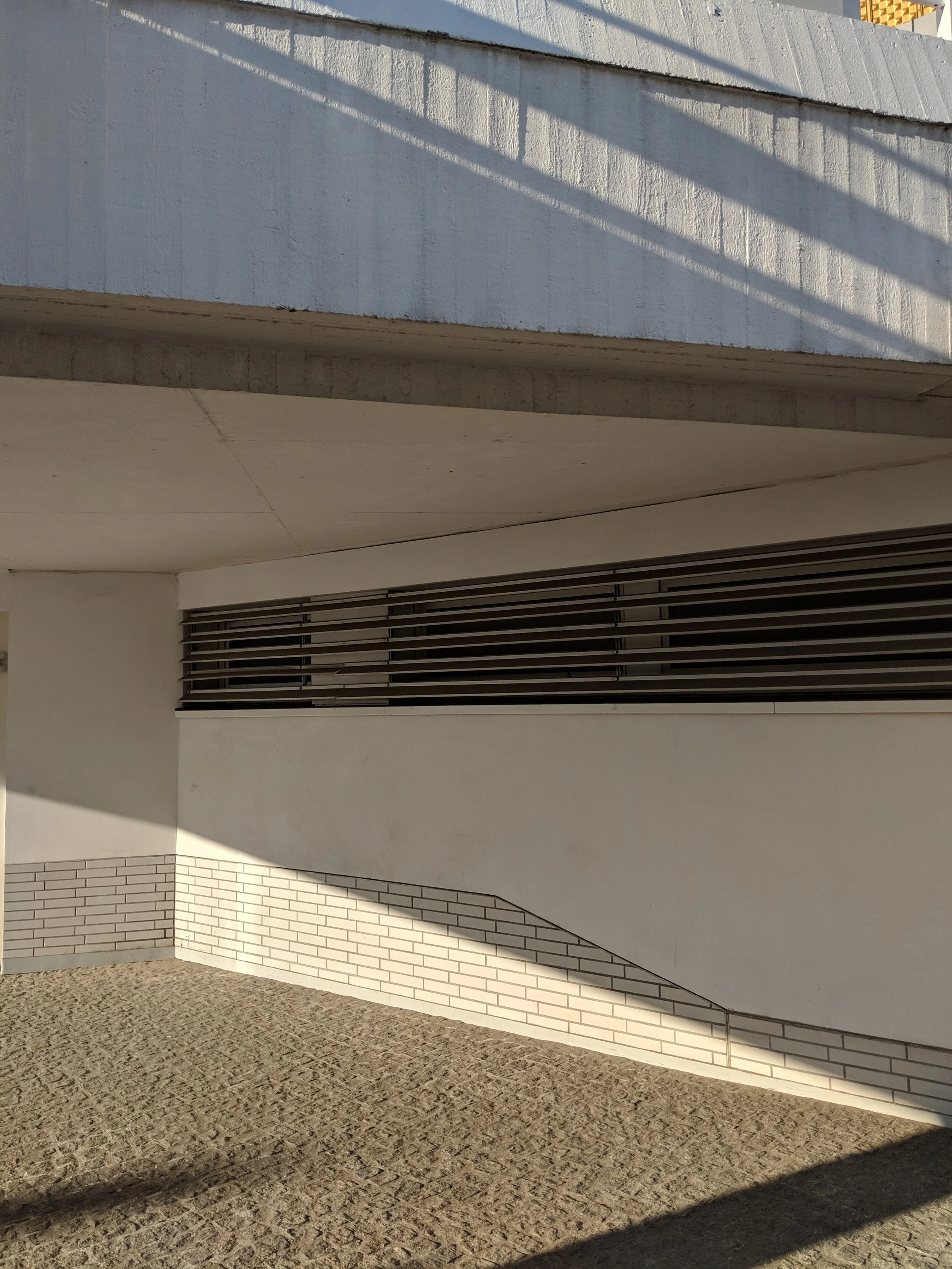 Modern building exterior with white walls, horizontal window blinds, and textured pavement, casting shadows.