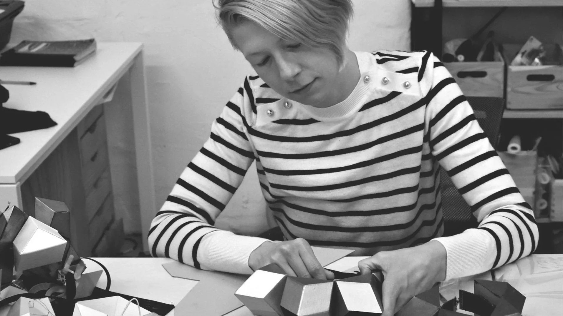 Black and white photo of a person with short, light-colored hair wearing a striped shirt with some decorative pearls, sitting at a desk with geometric paper crafts and a model of a geometric object on the desk, in a room with storage boxes in the background.