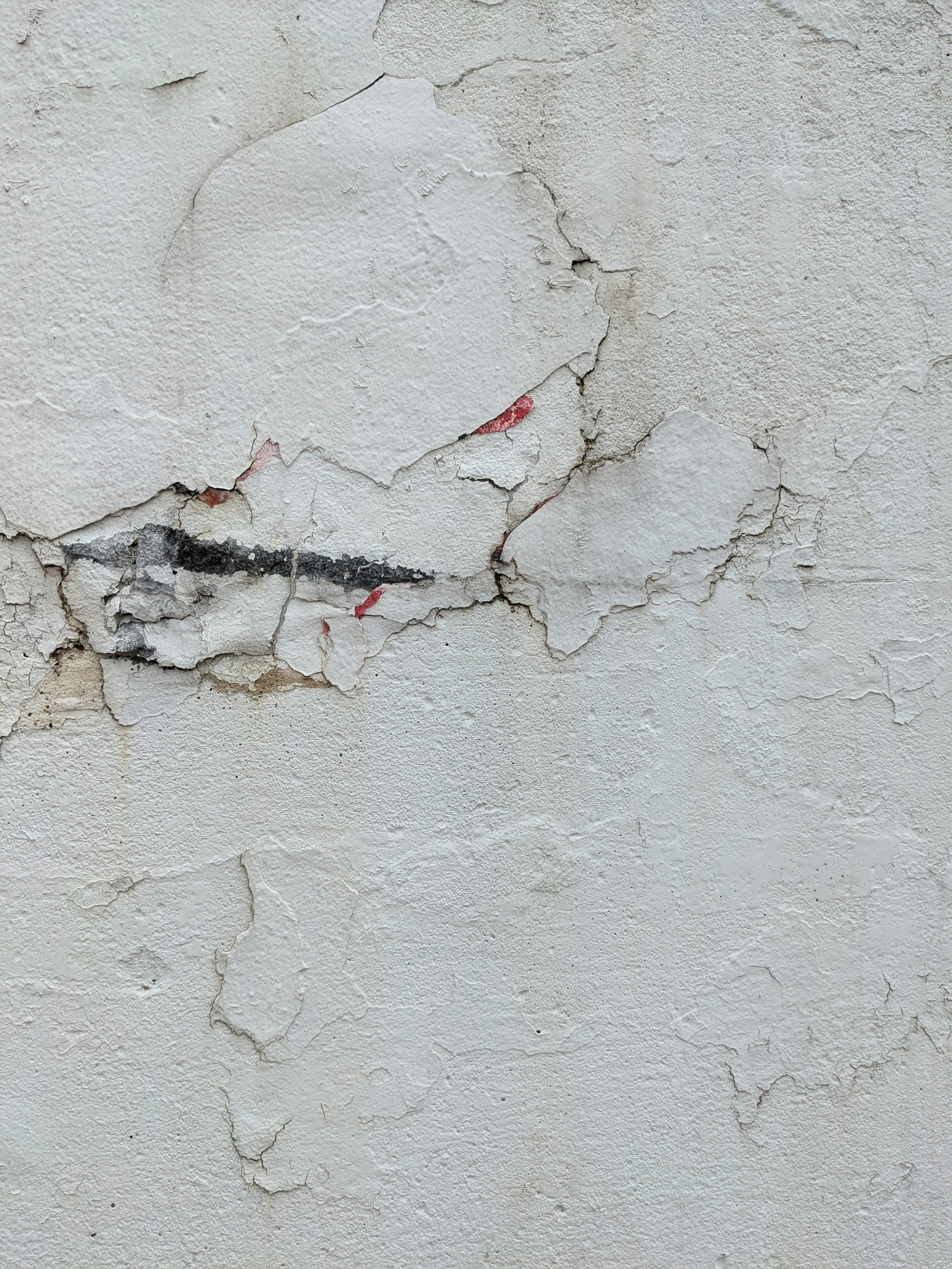 A close-up of a wall with peeling and cracked white paint, revealing underlying layers of different colors and textures.