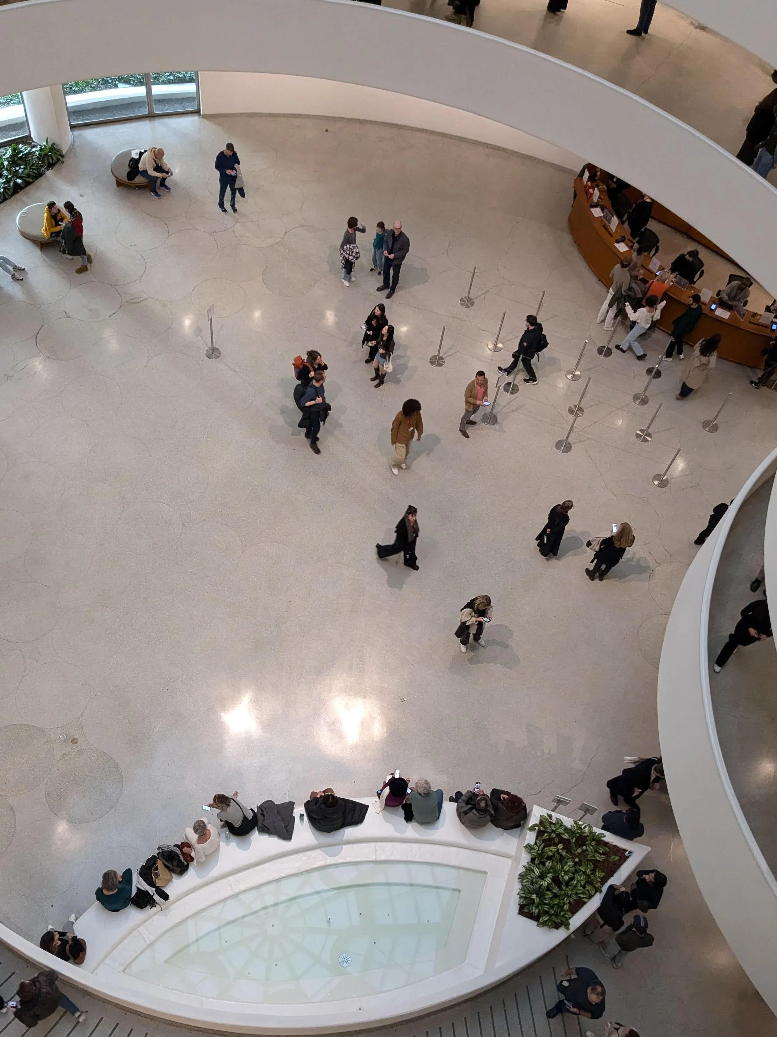 Interior view of a large, modern building with a circular mezzanine and spiral staircase. People are sitting, standing, and talking in the open, spacious atrium, with some walking or waiting in line at a service counter. There are also plants and lar