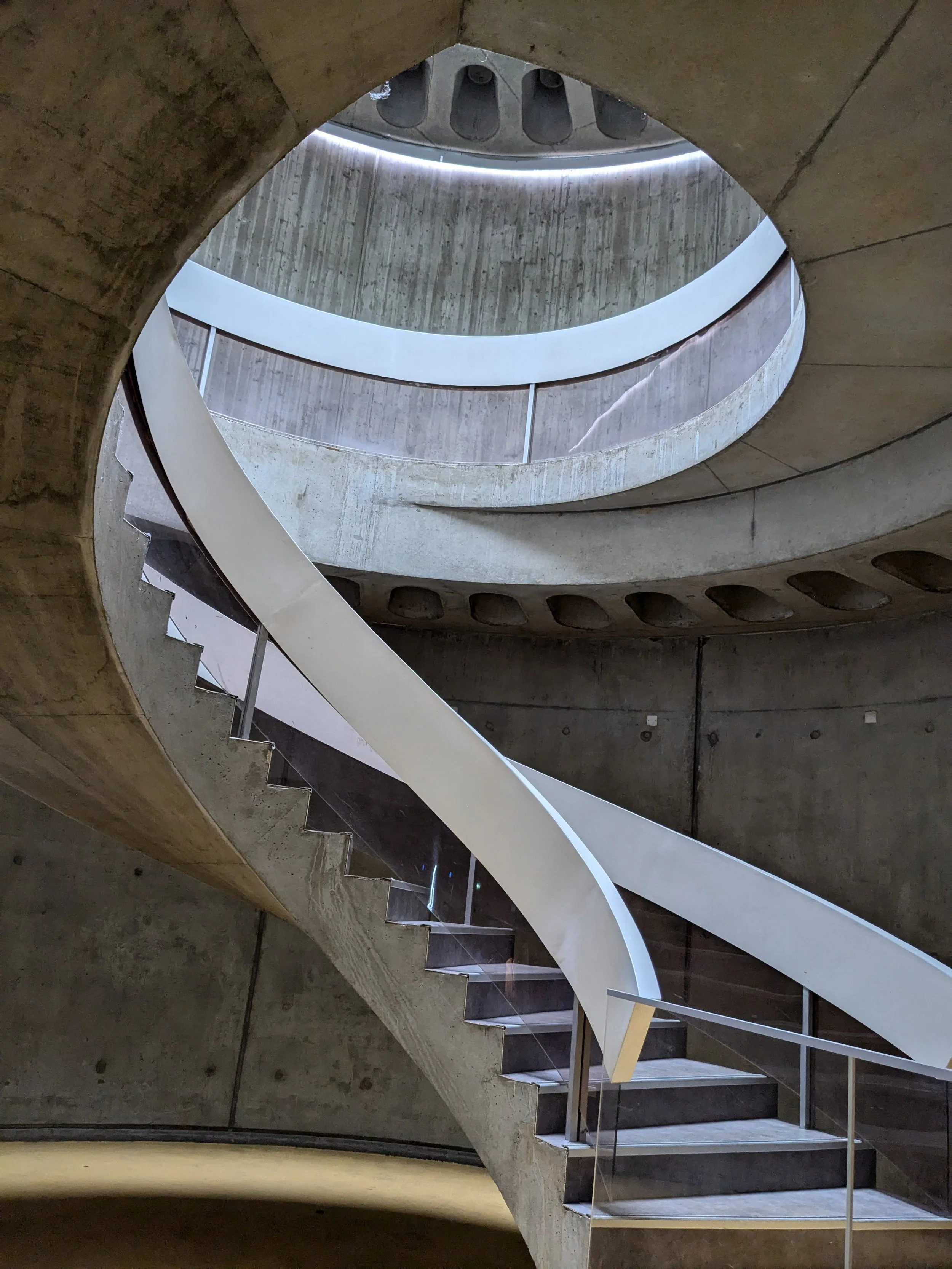 Inside view of a modern concrete spiral staircase with white railings and steps, and small windows on the wall at the bottom.