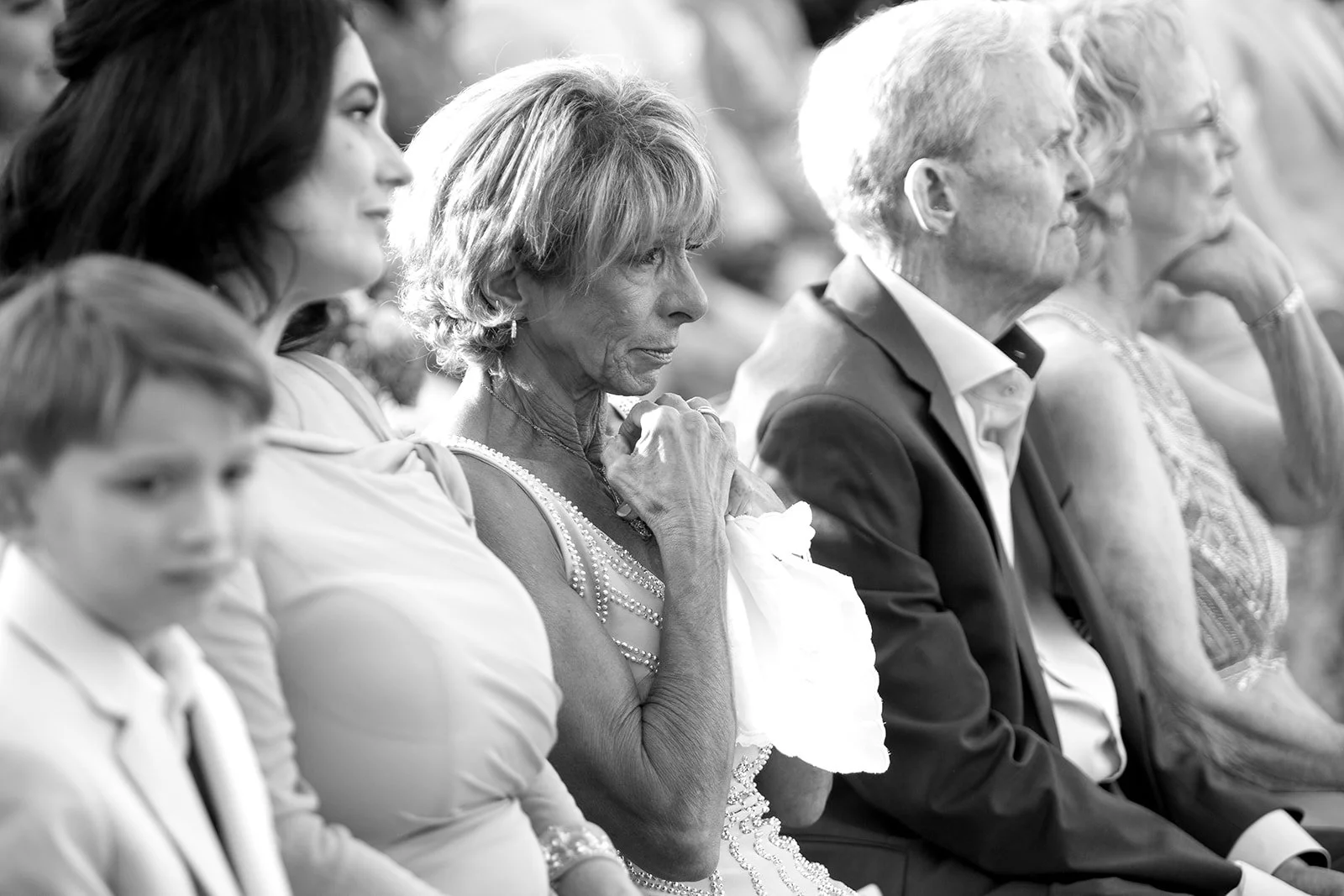 Black and white photo of a group of people sitting and listening at an event, including women, a man, and an elderly woman in focus.