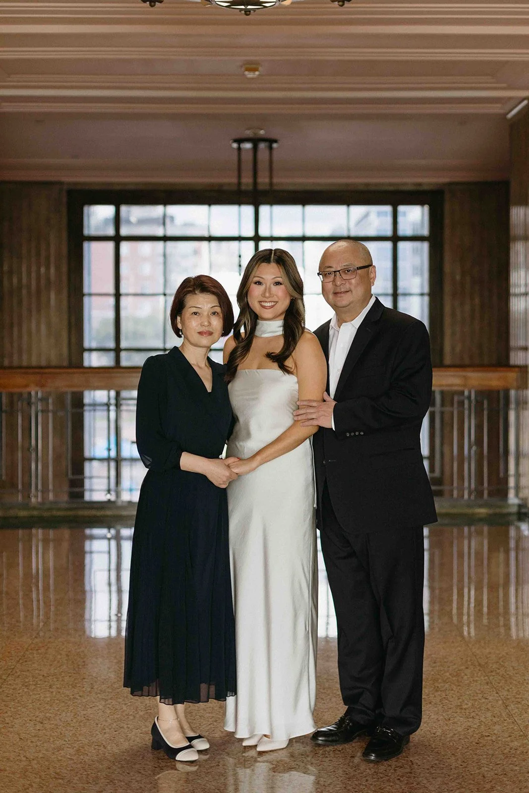 Three people standing close together in a well-lit indoor setting. The woman in the center is wearing a white dress, and the other two people, a woman and a man, are dressed in black. They are smiling and holding hands.