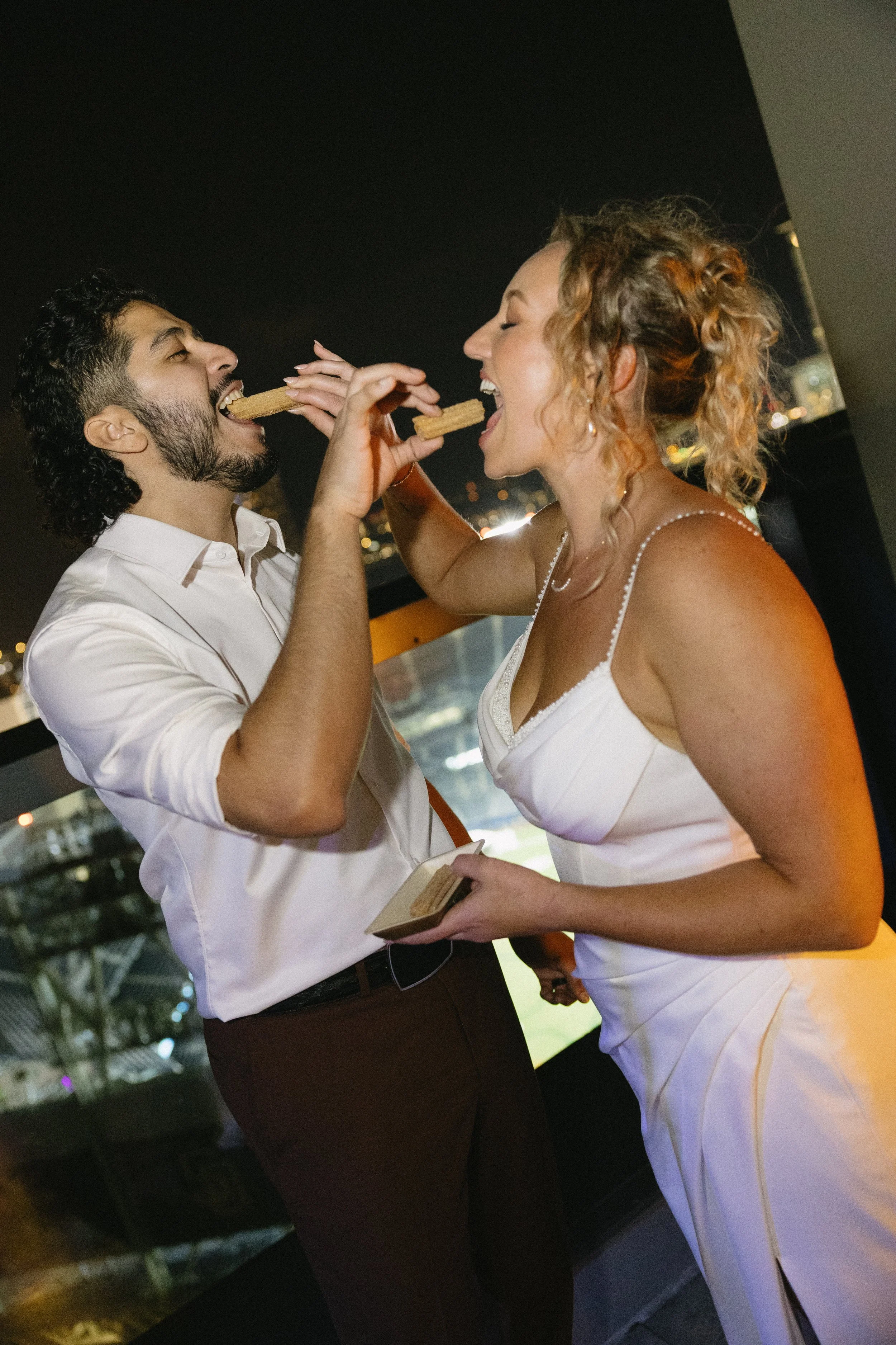 A man and a woman sharing a snack, with the woman holding a small plate, both smiling and laughing at night in an urban setting.