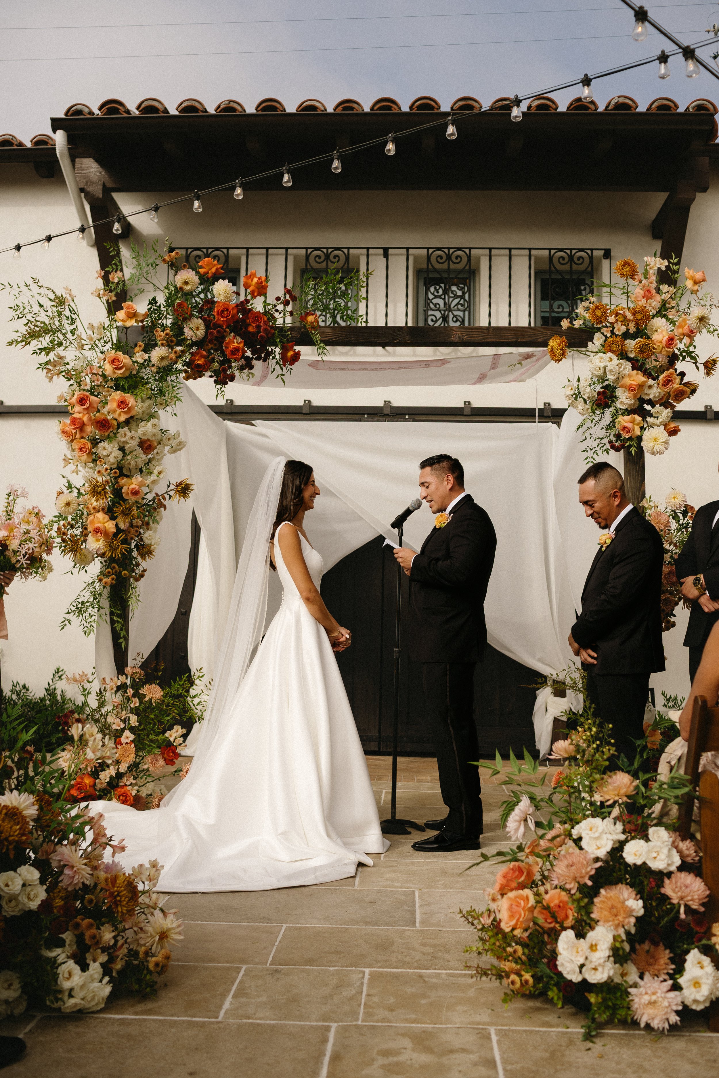Bride and groom exchanging vows during outdoor wedding ceremony, surrounded by floral arrangements, with officiant reading from paper, guests seated nearby.