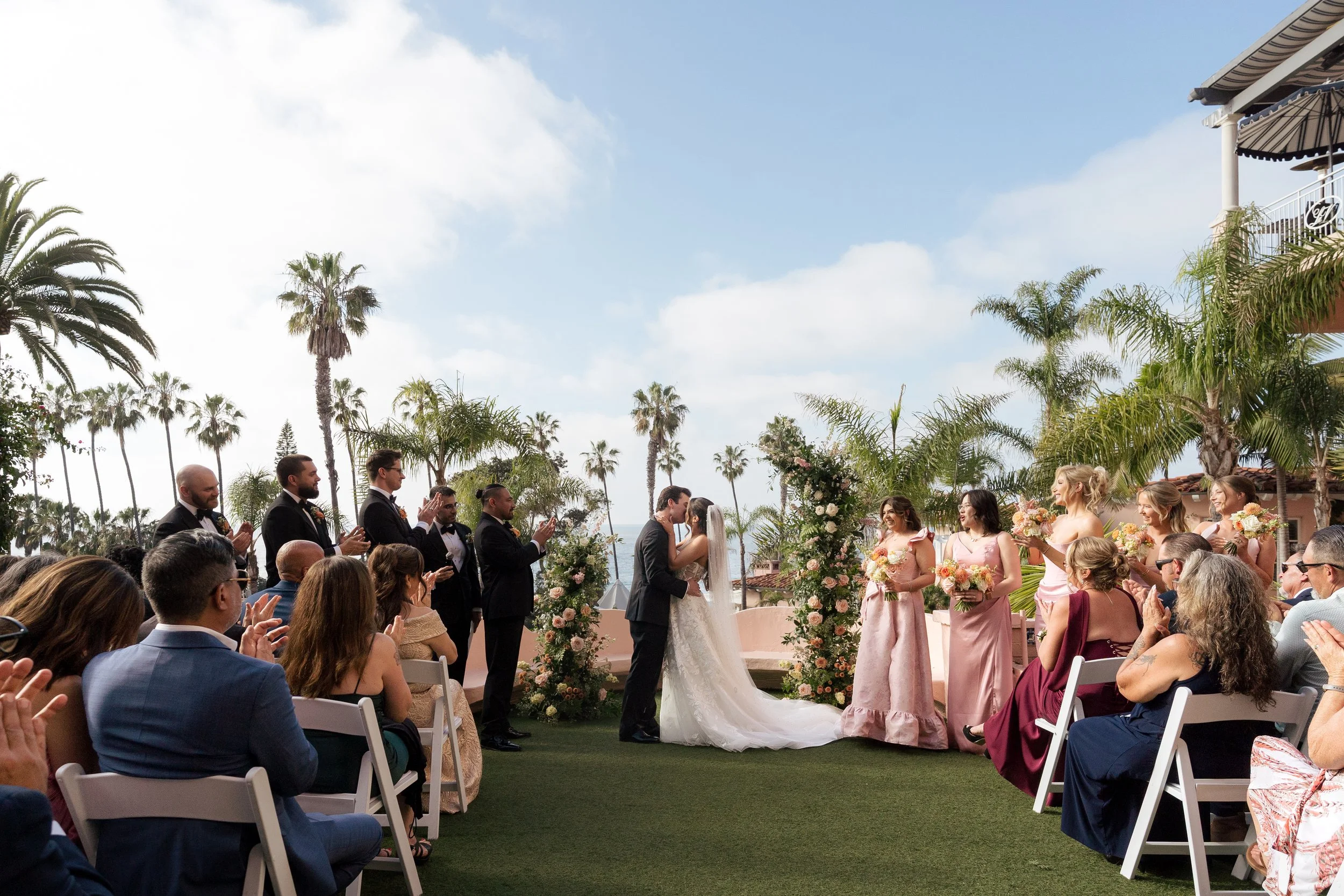 A wedding ceremony outdoors with a bride and groom exchanging vows, surrounded by bridesmaids and groomsmen, with guests seated and clapping, palm trees, and a blue sky with some clouds.