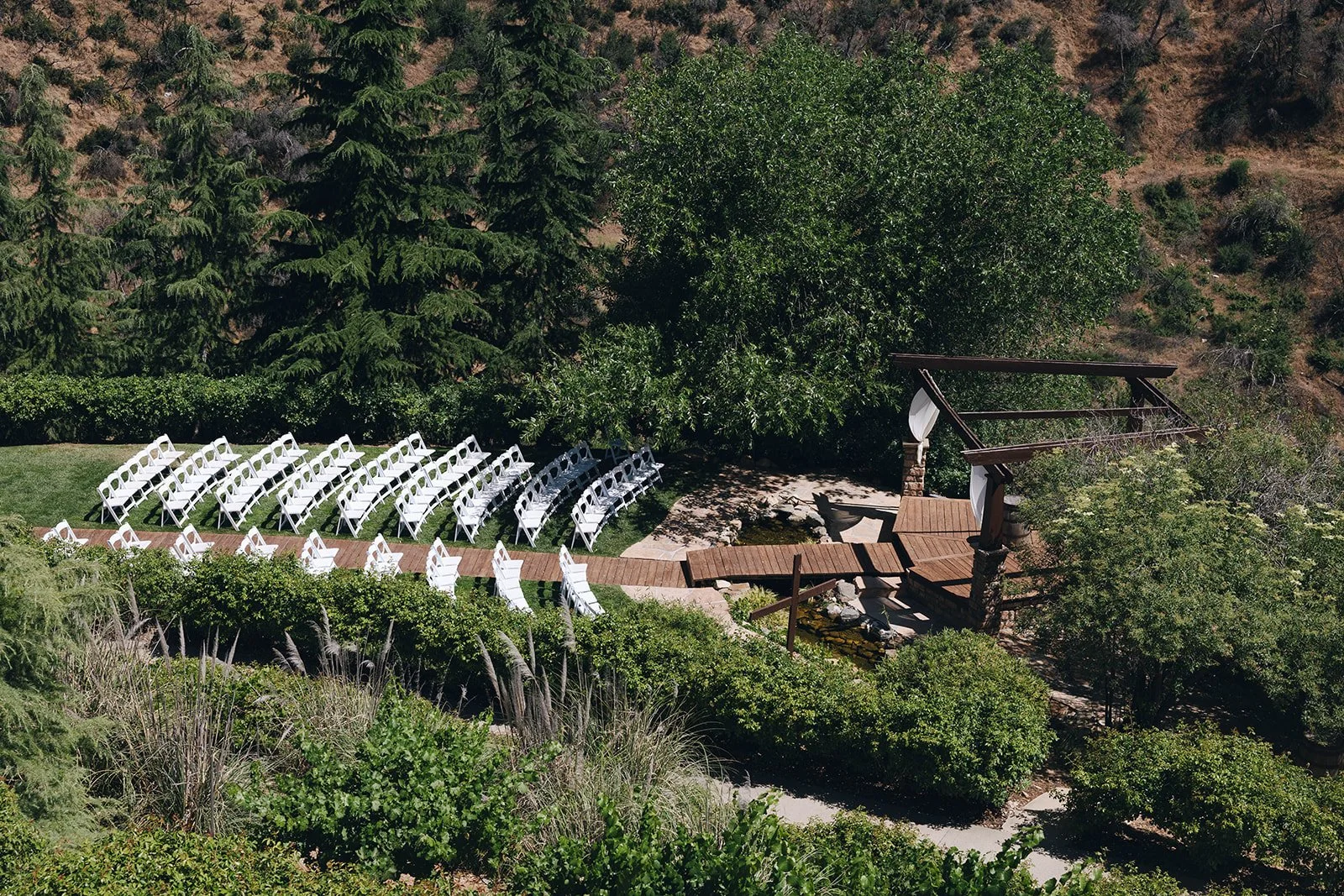 Outdoor wedding ceremony setup with white chairs arranged on grass facing a decorated wooden arch, surrounded by lush green trees and shrubs in a garden area.