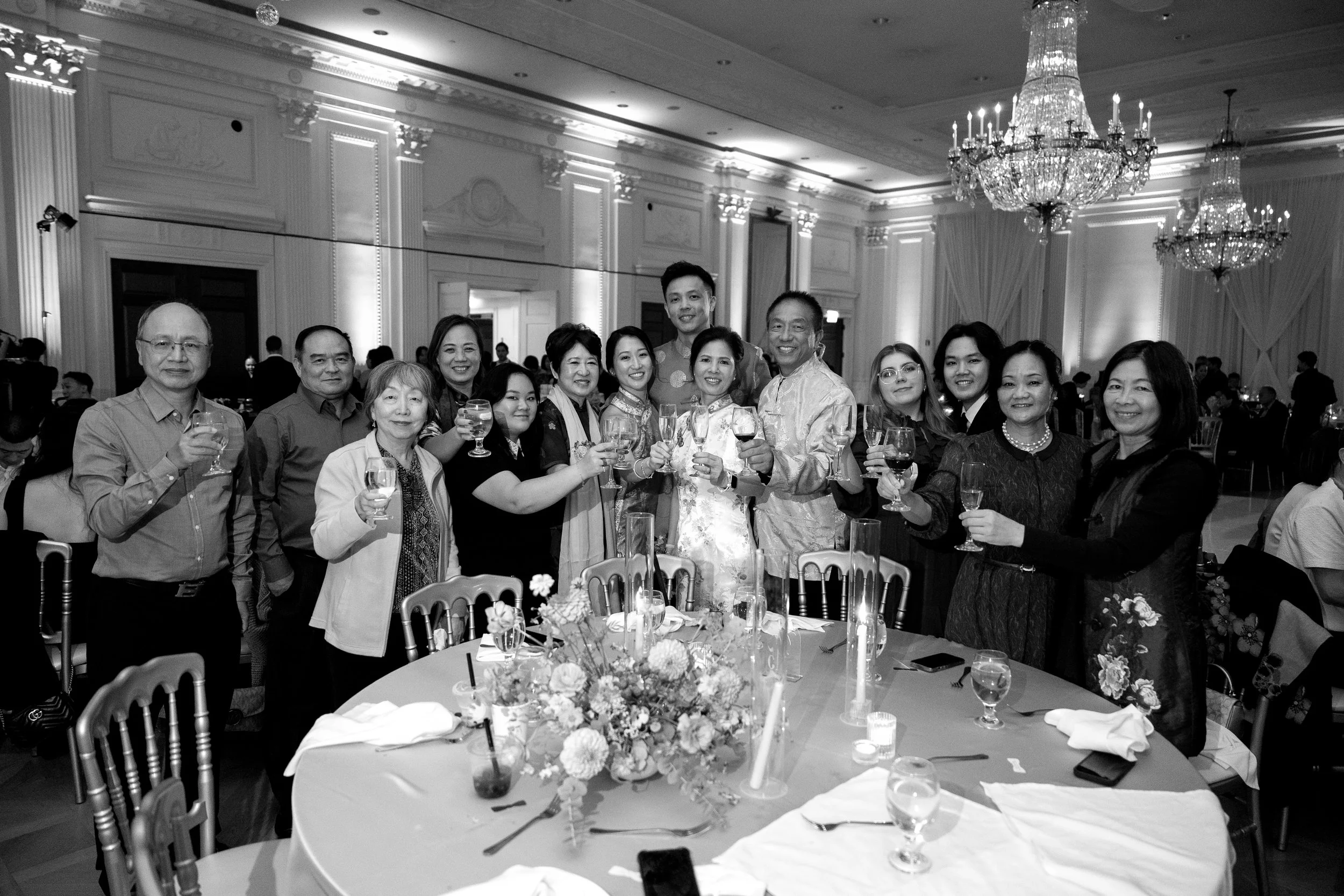 A group of people dressed formally, celebrating at a banquet table with glasses of wine, in an elegant hall with chandeliers and floral centerpiece.