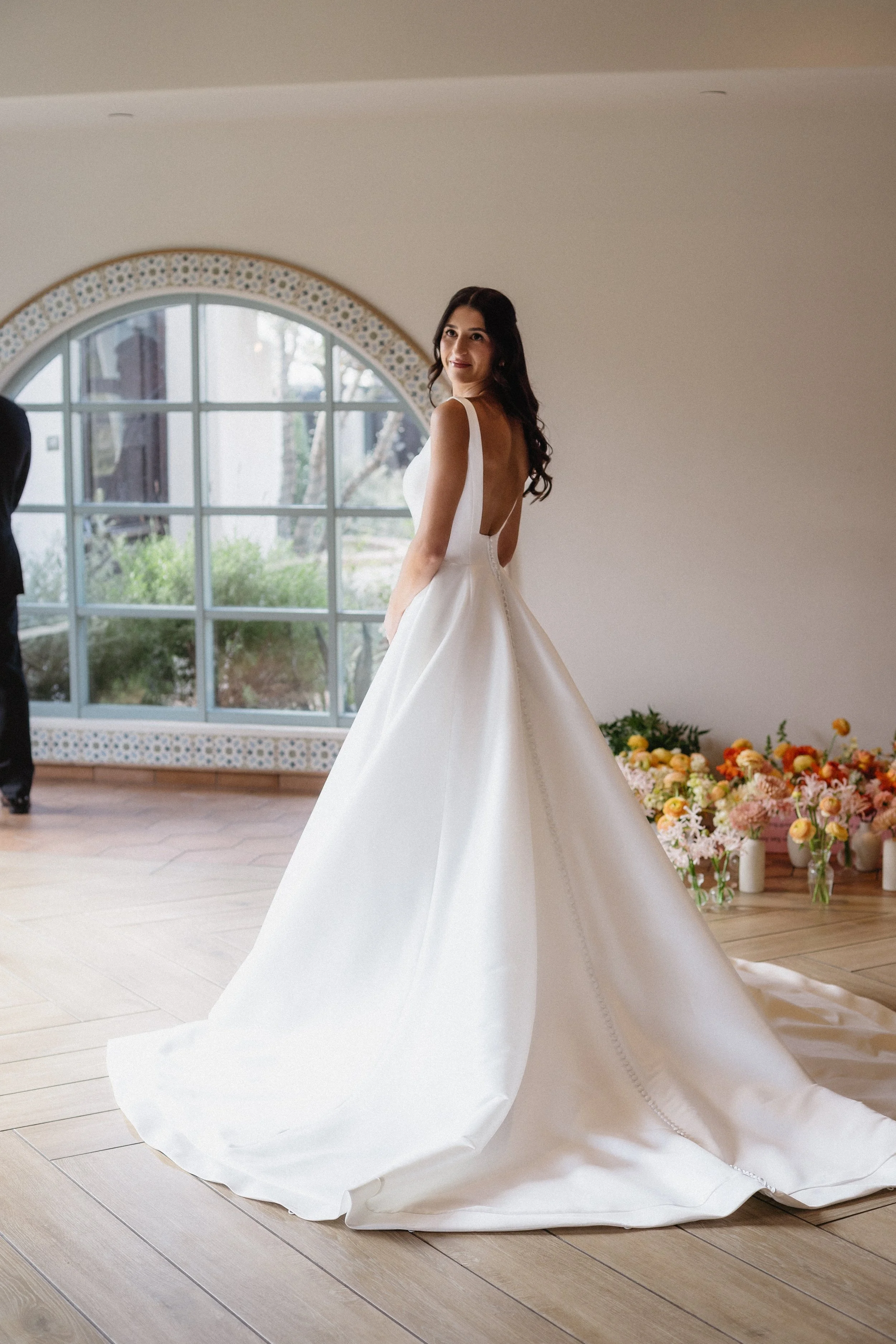 A woman in a white wedding gown standing indoors near a large arched window with a garden view and floral decorations on the floor.