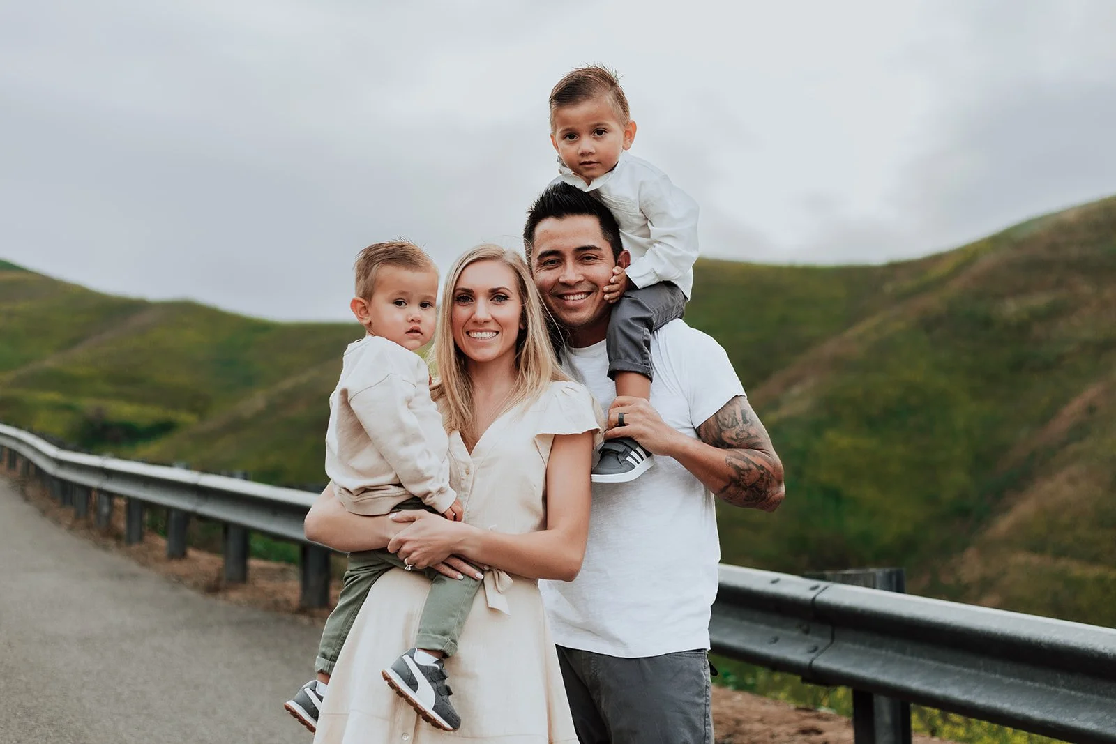 Family of four standing outdoors on a cloudy day, with green hills in the background. The mother has blonde hair and is smiling, holding a young boy. The father, with tattoos and dark hair, is smiling and carrying a slightly older boy on his shoulders.