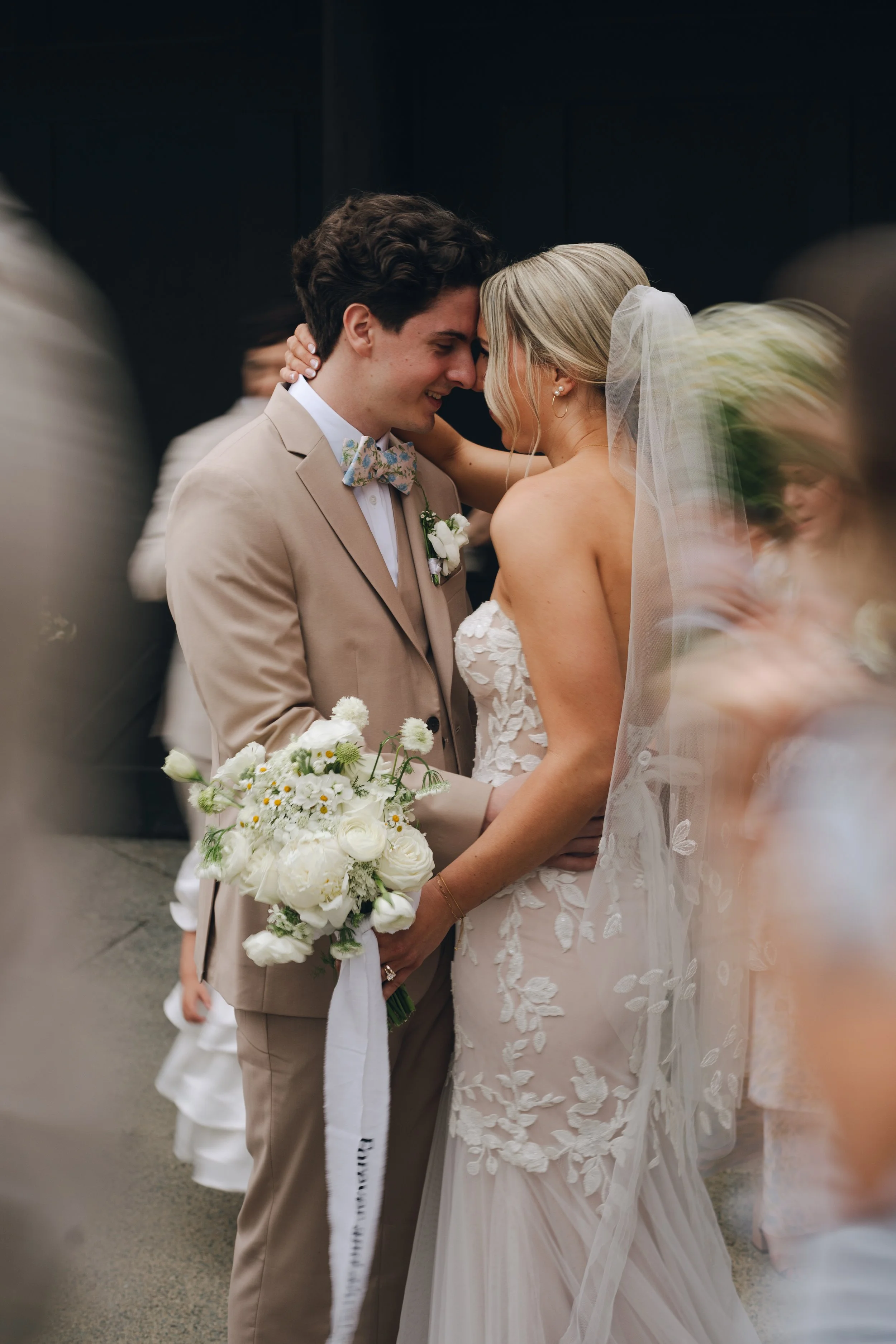 A bride and groom share a close, intimate moment during their wedding ceremony, smiling with their foreheads touching, surrounded by blurred guests.