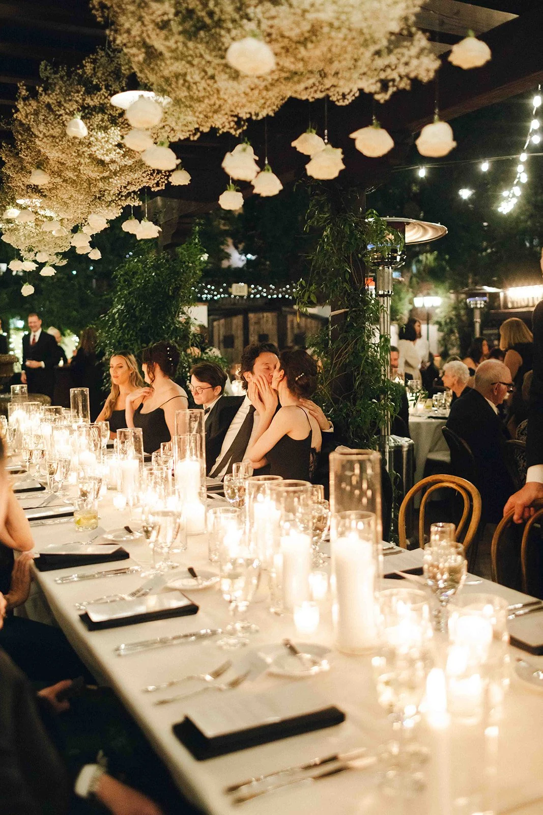 People sitting at a decorated dinner table under hanging floral arrangements with candles and string lights in the evening