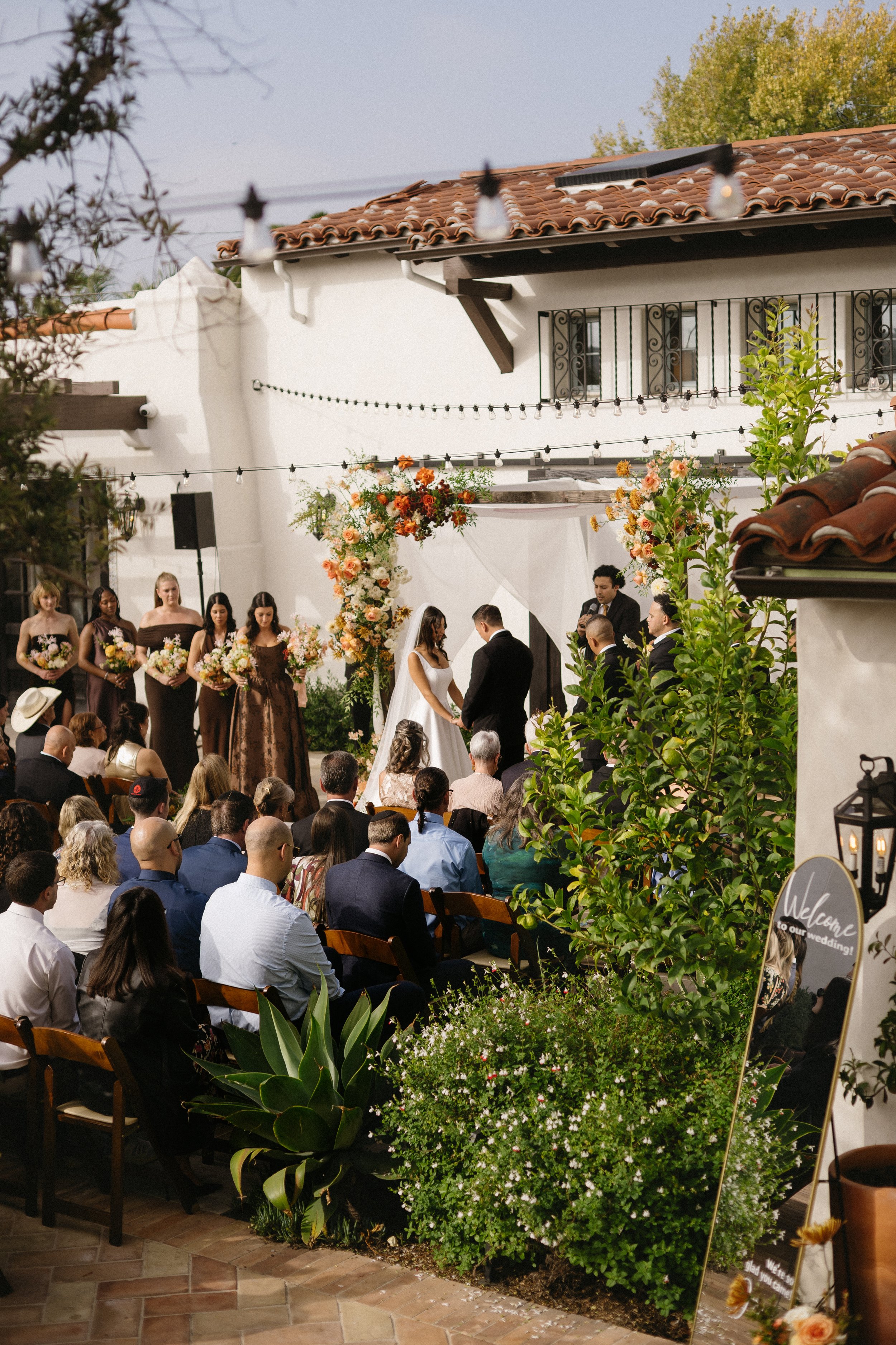 Outdoor wedding ceremony with a couple exchanging vows under a floral arch, surrounded by guests and bridesmaids in brown and mauve dresses, with lush greenery and a welcoming sign.