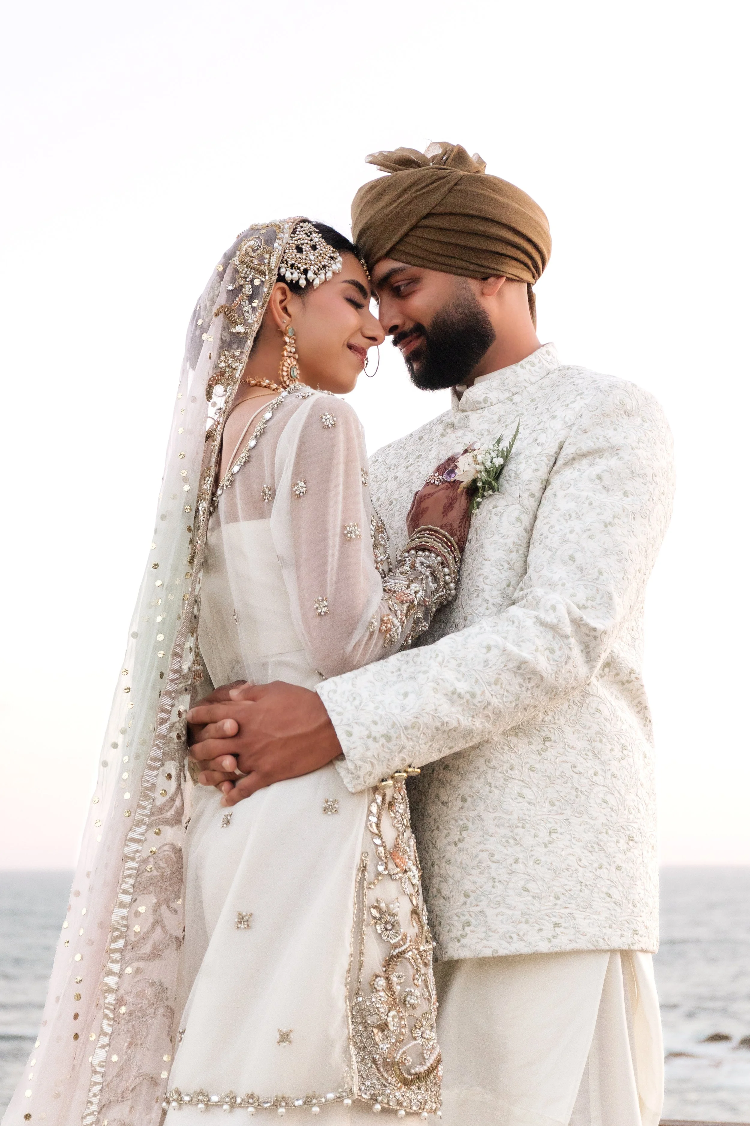 A couple dressed in traditional Indian wedding attire holding hands, with their foreheads touching, by the beach.