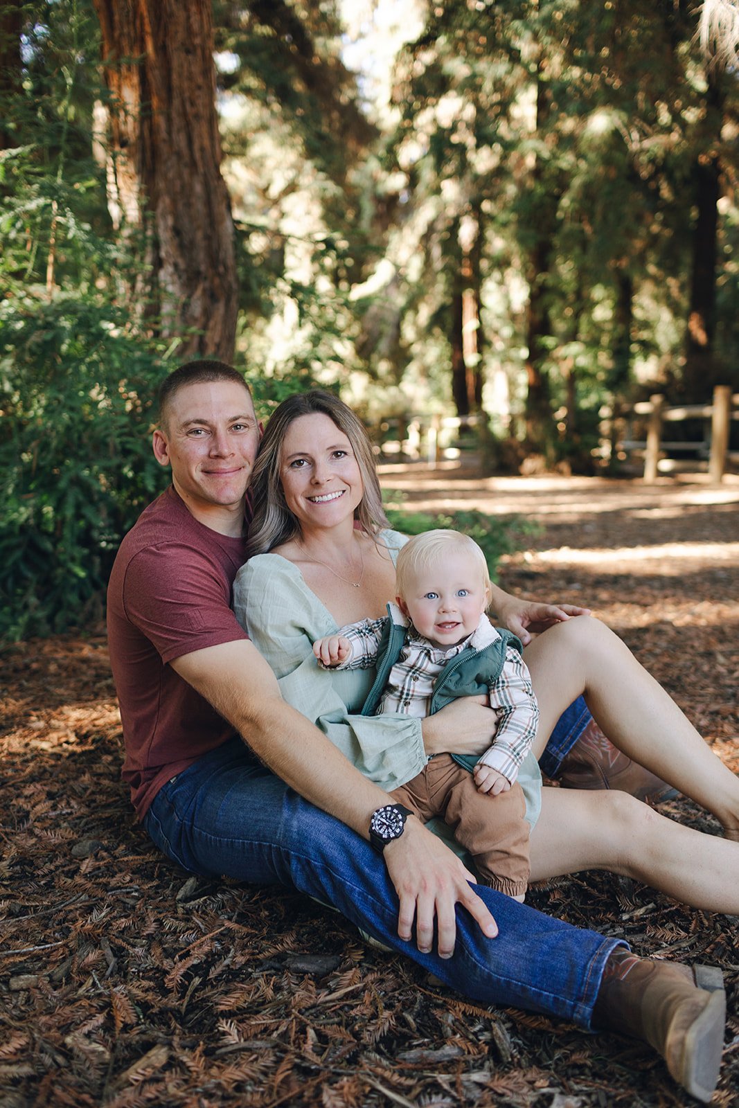A family of three sitting on the forest floor, smiling at the camera.